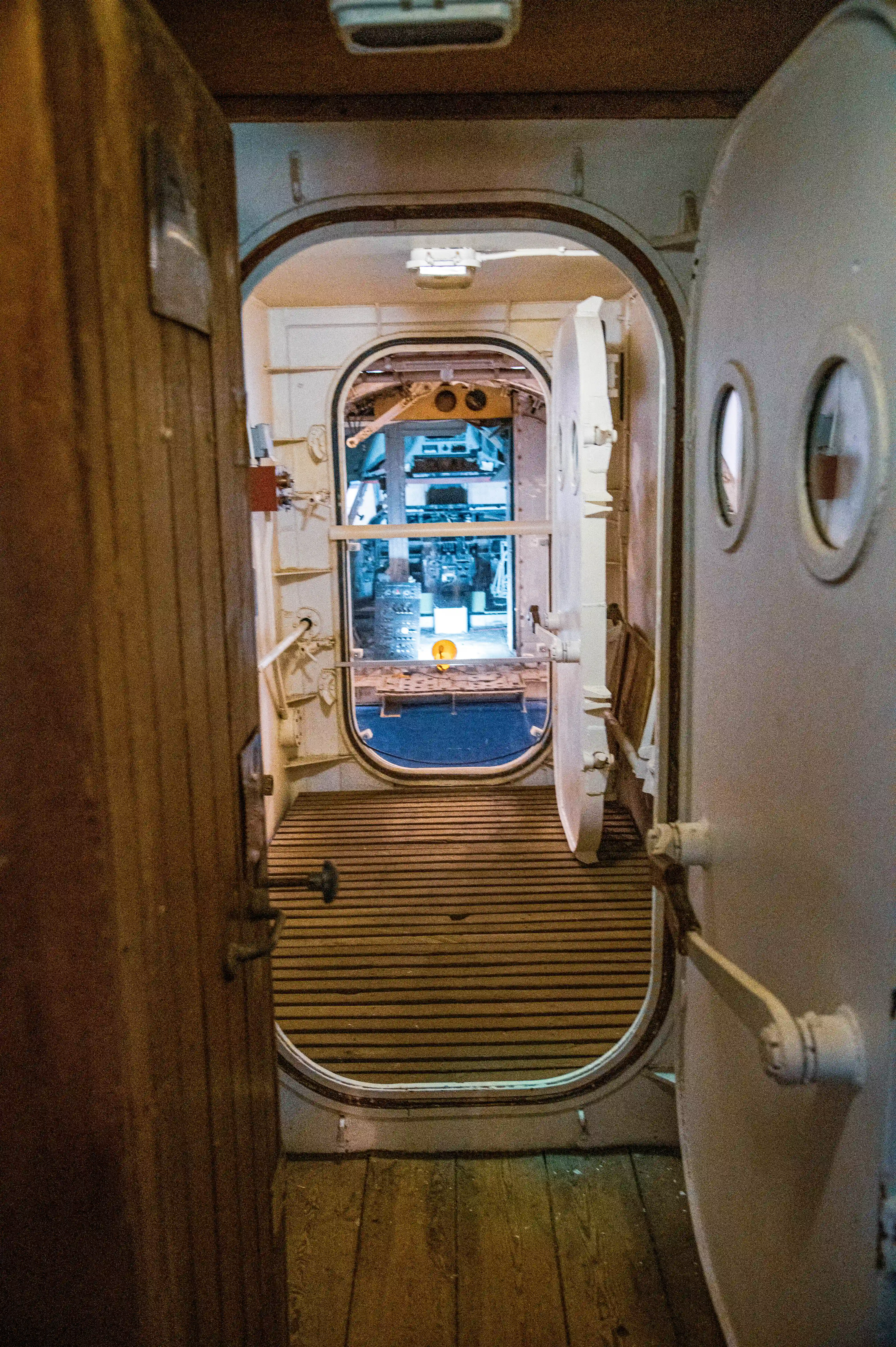 A view of the interior of an aircraft fuselage, showcasing the cockpit area with visible instrument panels, control yokes, and seating. The fuselage features metal framing, rivets, and a circular door opening, with a red ladder leading up to the entrance. The background includes hangar walls and lighting.