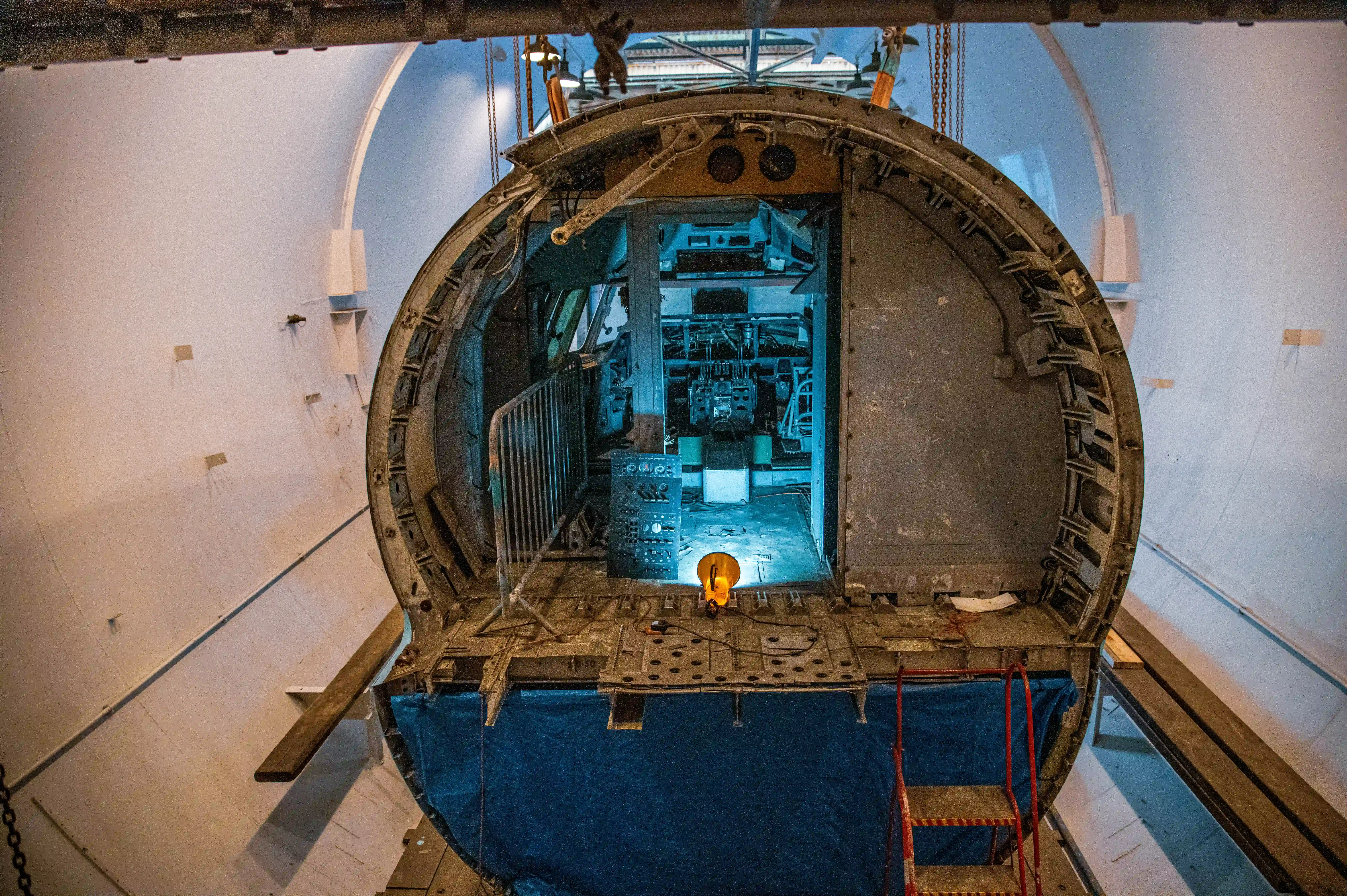 Interior view of a air-lock showing an open hatchway. The foreground features wooden flooring and a partially open wooden door on the left. Ahead, a curved metal doorway leads into a well-lit compartment with blue flooring and visible machinery.