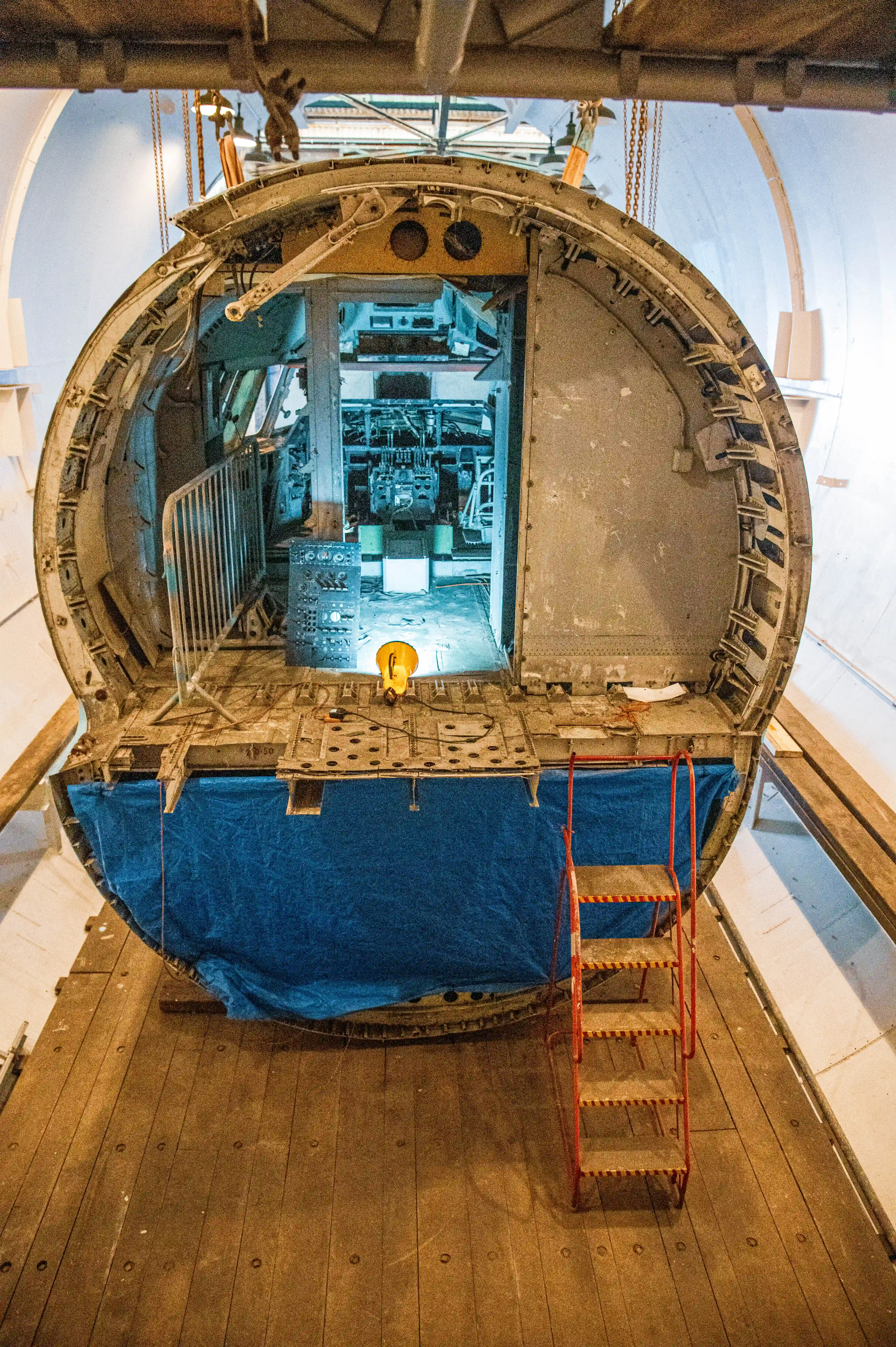 A view of the interior of an aircraft fuselage, showcasing the cockpit area with visible instrument panels, control yokes, and seating. The fuselage features metal framing, rivets, and a circular door opening, with a red ladder leading up to the entrance. The background includes hangar walls and lighting.