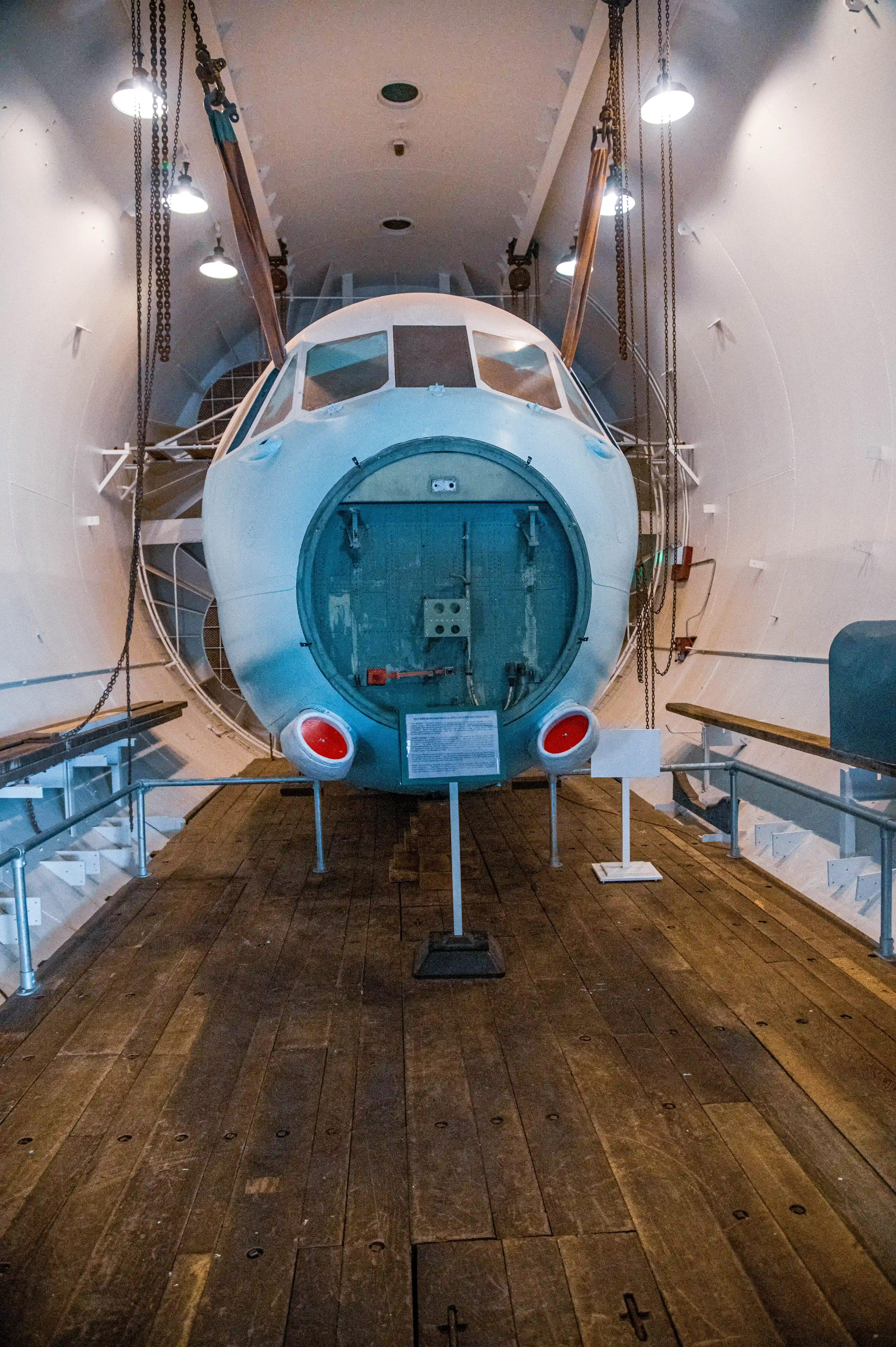 A view of the interior of an aircraft fuselage, showcasing the cockpit area with visible instrument panels, control yokes, and seating. The fuselage features metal framing, rivets, and a circular door opening, with a red ladder leading up to the entrance. The background includes hangar walls and lighting.