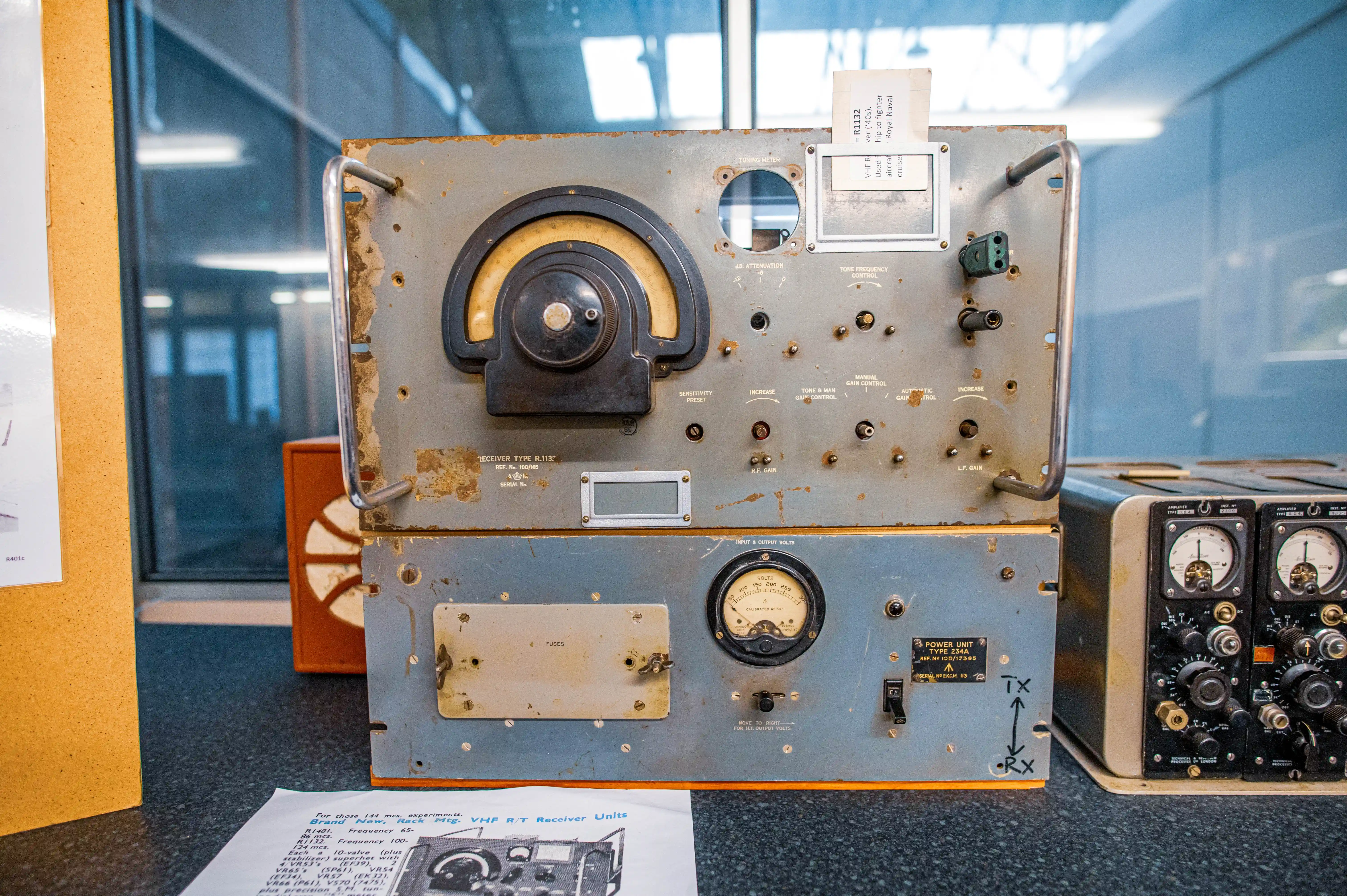 Two vintage radio receiver units are displayed side-by-side. The top unit has a large tuning dial and numerous smaller control knobs. The bottom unit features a large VU meter and a fused power switch. Both units appear to be constructed of metal with a grey finish and show signs of age.