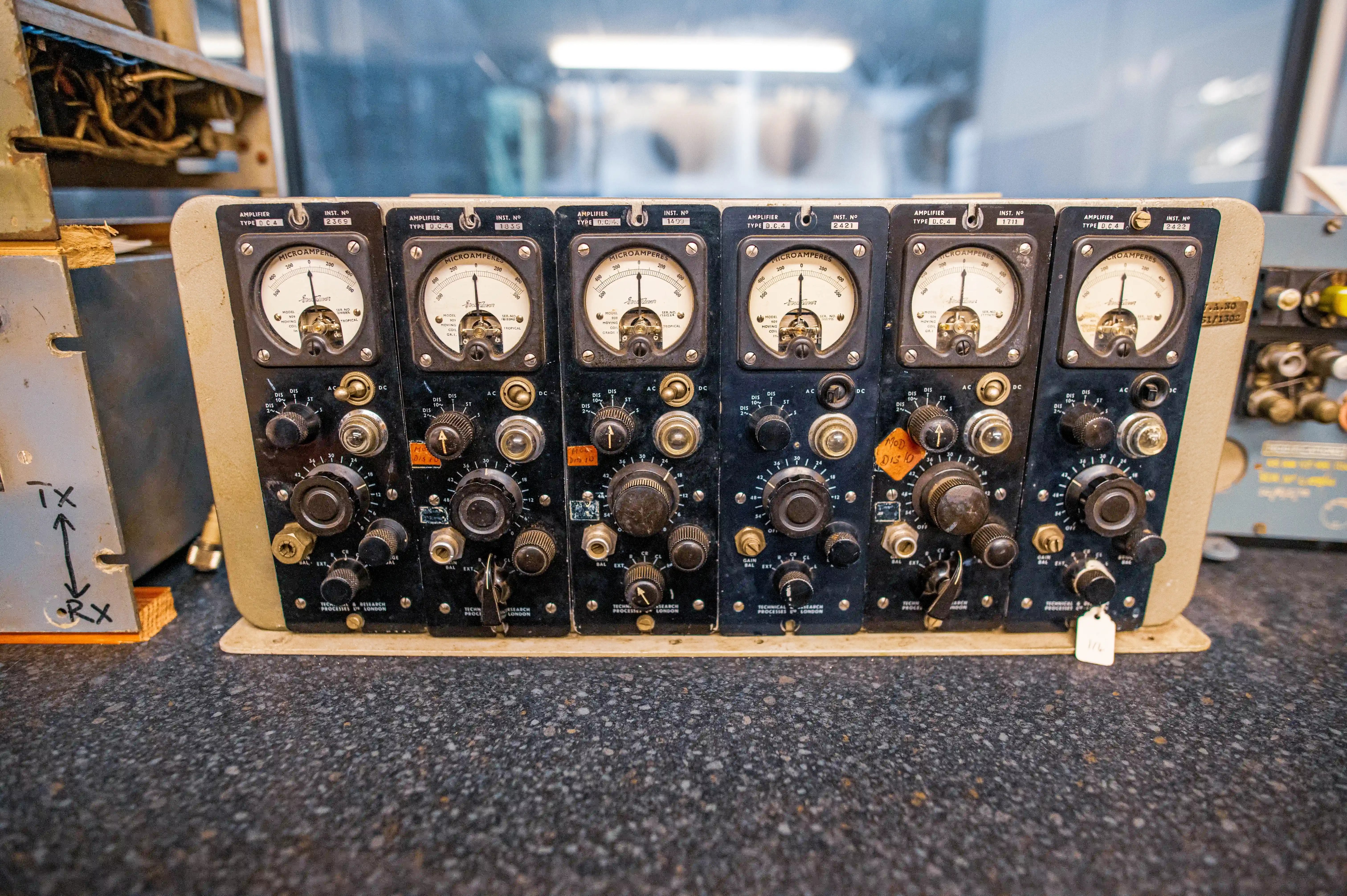 A close-up frontal view of a row of six vintage electrical amplifier units. Each unit features a central analogue microammeter displaying readings from 0 to 500, a type designation of AMPLIFIER TYPE D.C.4, and an INST. NO. serial number. Below the meters are arrays of black rotary knobs, toggle switches, and circular dials with various settings. Some units have small orange labels with handwritten text such as MOD. DIS. 10. To the far left, a separate metal unit displays handwritten TX with an upward arrow and RX with a downward arrow. The equipment sits on a dark, speckled surface, with blurred laboratory equipment visible in the background.
