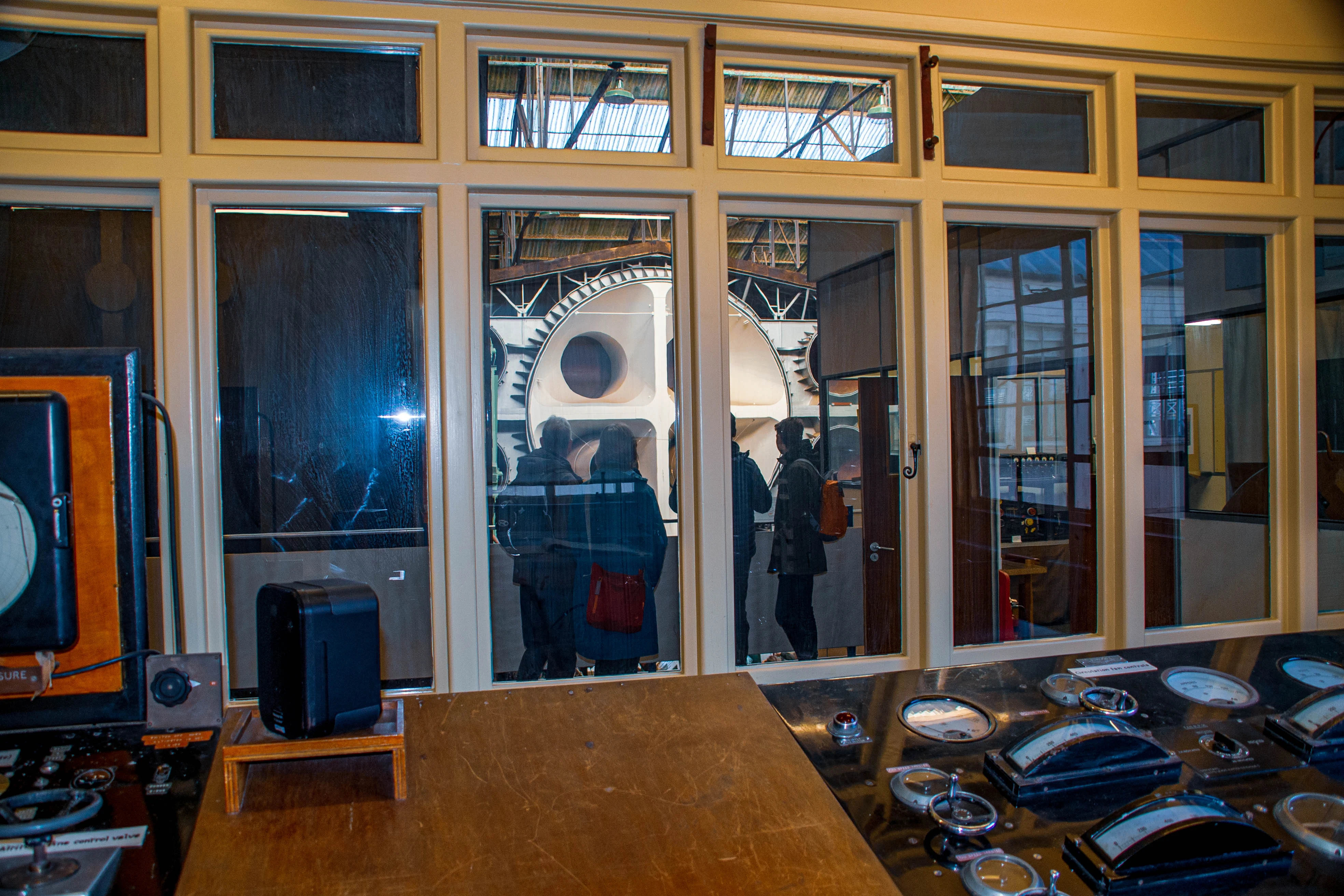 View from inside a control room with a panel of gauges and levers, looking out through large windows to an industrial space with large machinery and several people observing.
