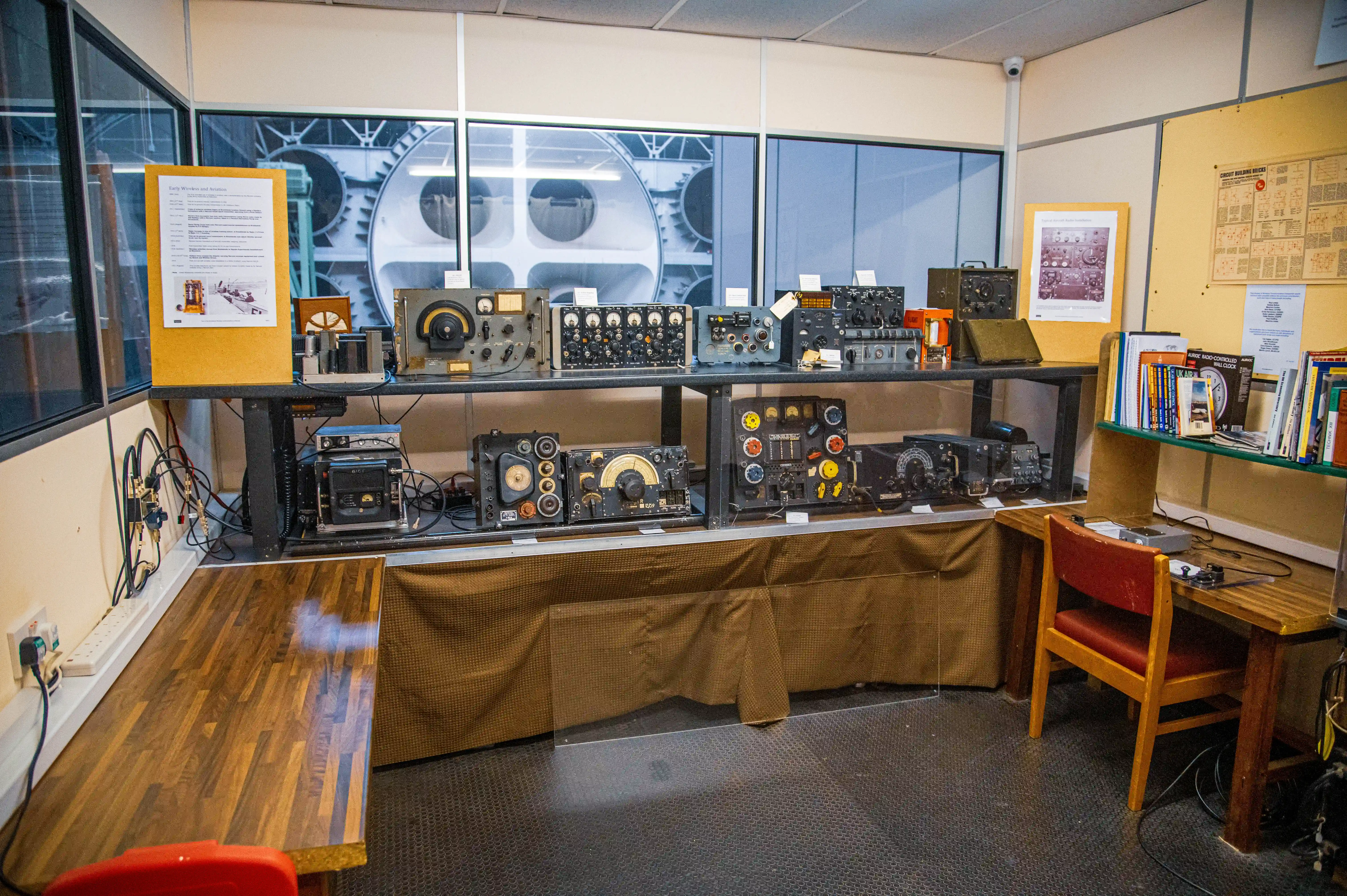 A museum display of vintage radio equipment, featuring numerous analogue devices with dials and knobs on shelves, behind a protective barrier. A large cogwheel is visible through a window in the background. A wooden desk with books and a chair is on the right.