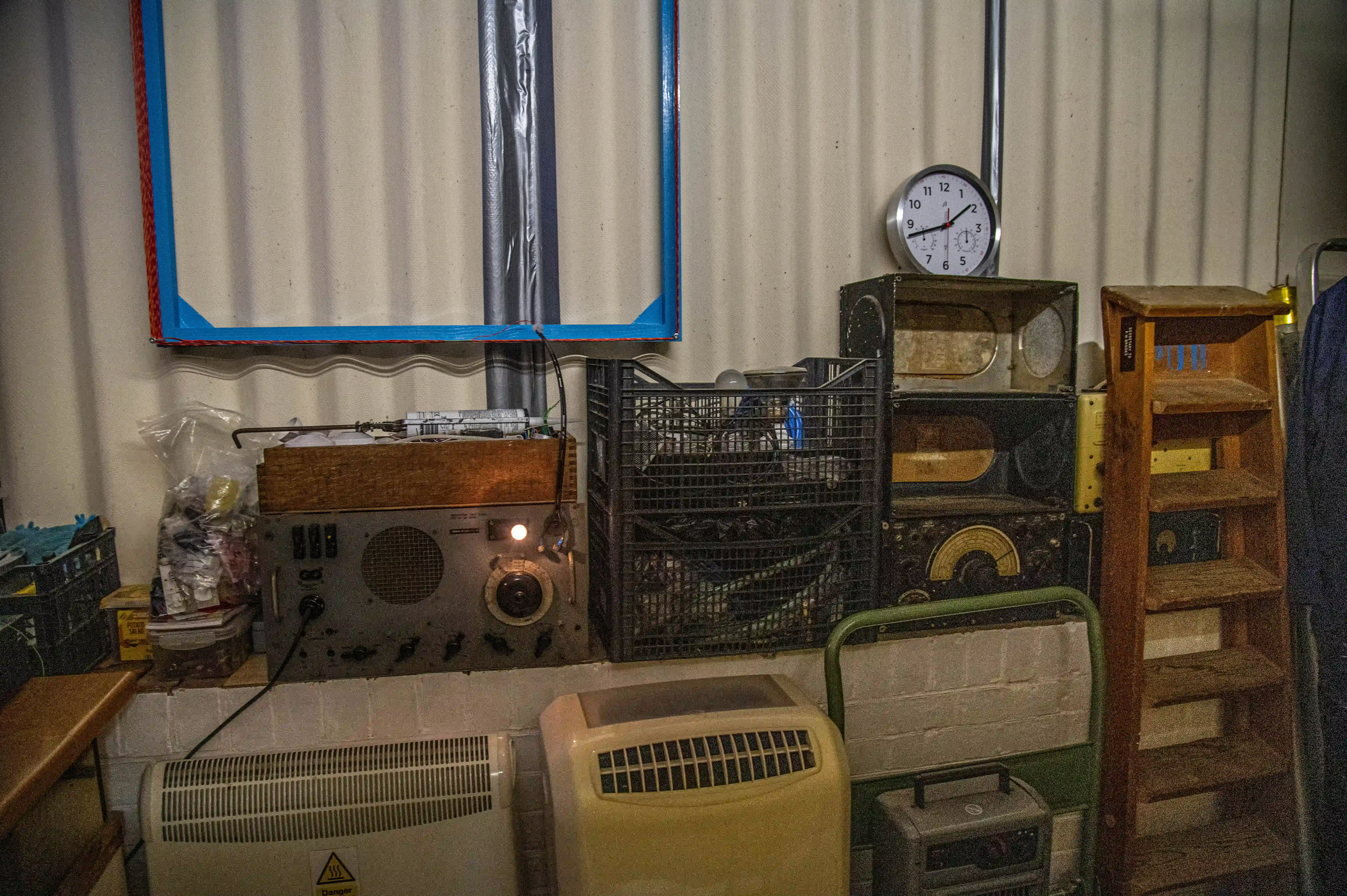 A museum display of vintage radio equipment, featuring numerous analogue devices with dials and knobs on shelves, behind a protective barrier. A large cogwheel is visible through a window in the background. A wooden desk with books and a chair is on the right.