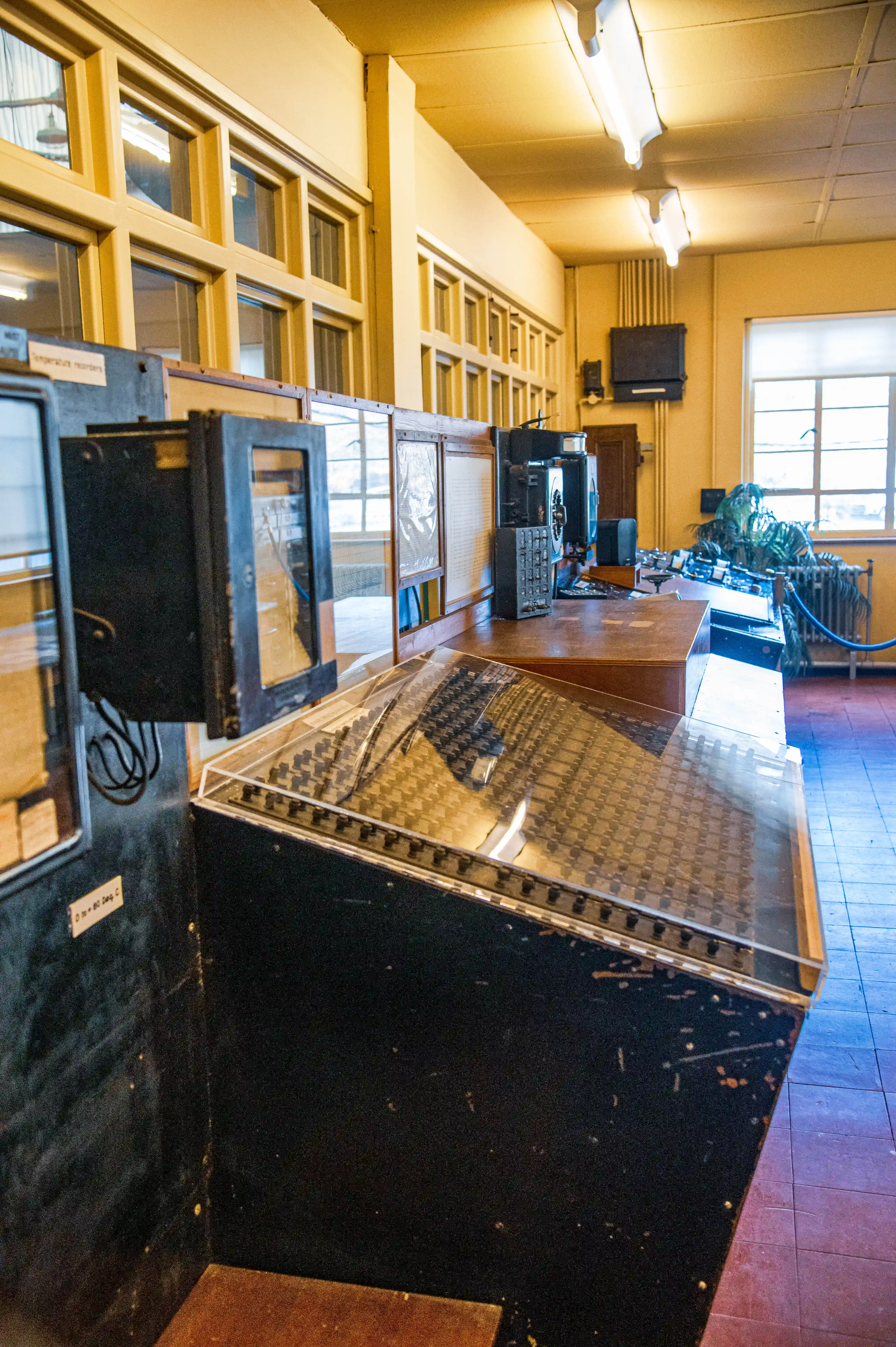A view inside a vintage control room featuring a large, dark-coloured console with a textured surface beneath a clear plastic cover. Several older electronic devices and wall-mounted recorders are visible, along with a bank of windows in the background.