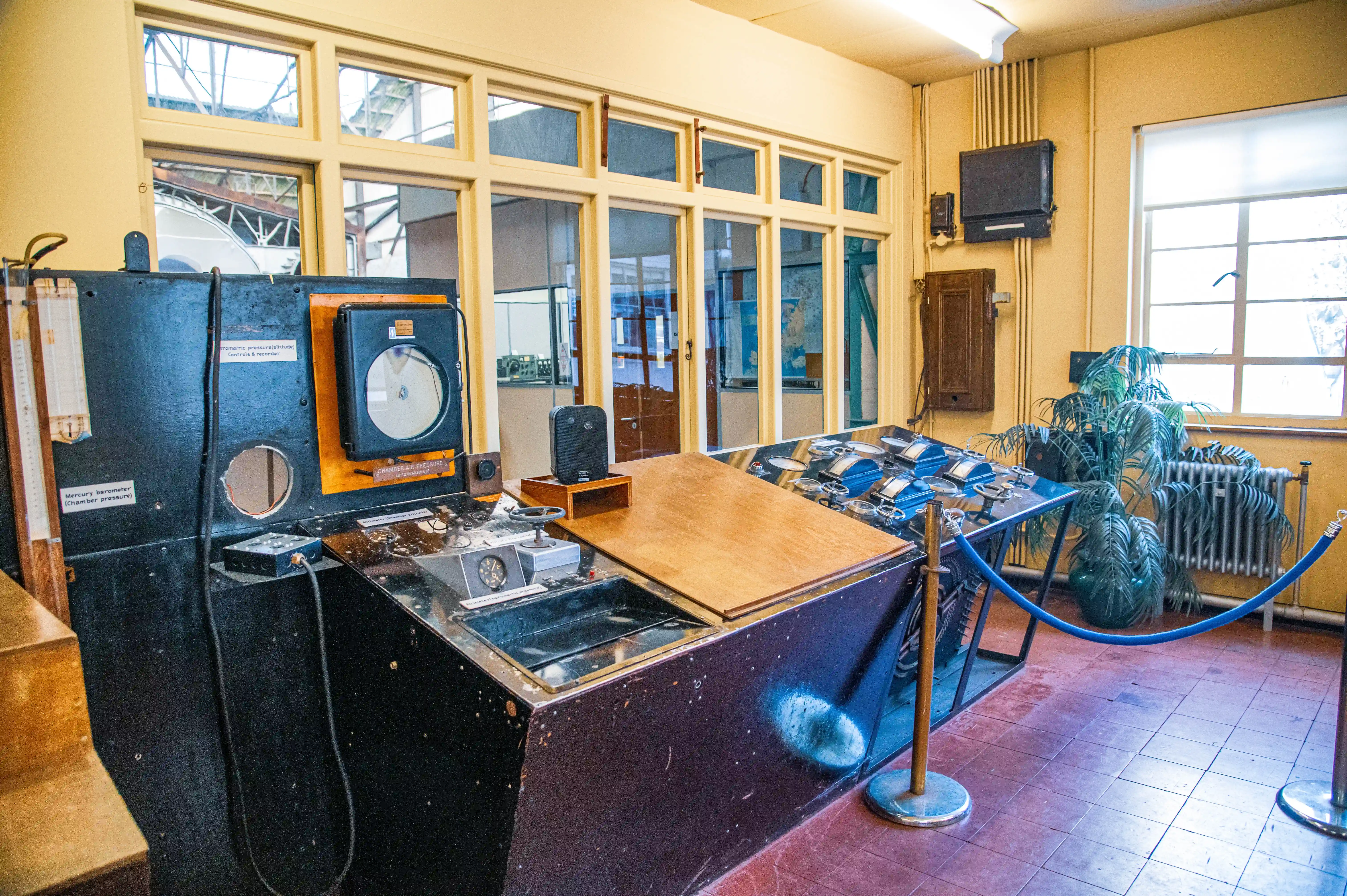 A view inside a vintage control room featuring a large, dark-coloured console with a textured surface beneath a clear plastic cover. Several older electronic devices and wall-mounted recorders are visible, along with a bank of windows in the background.