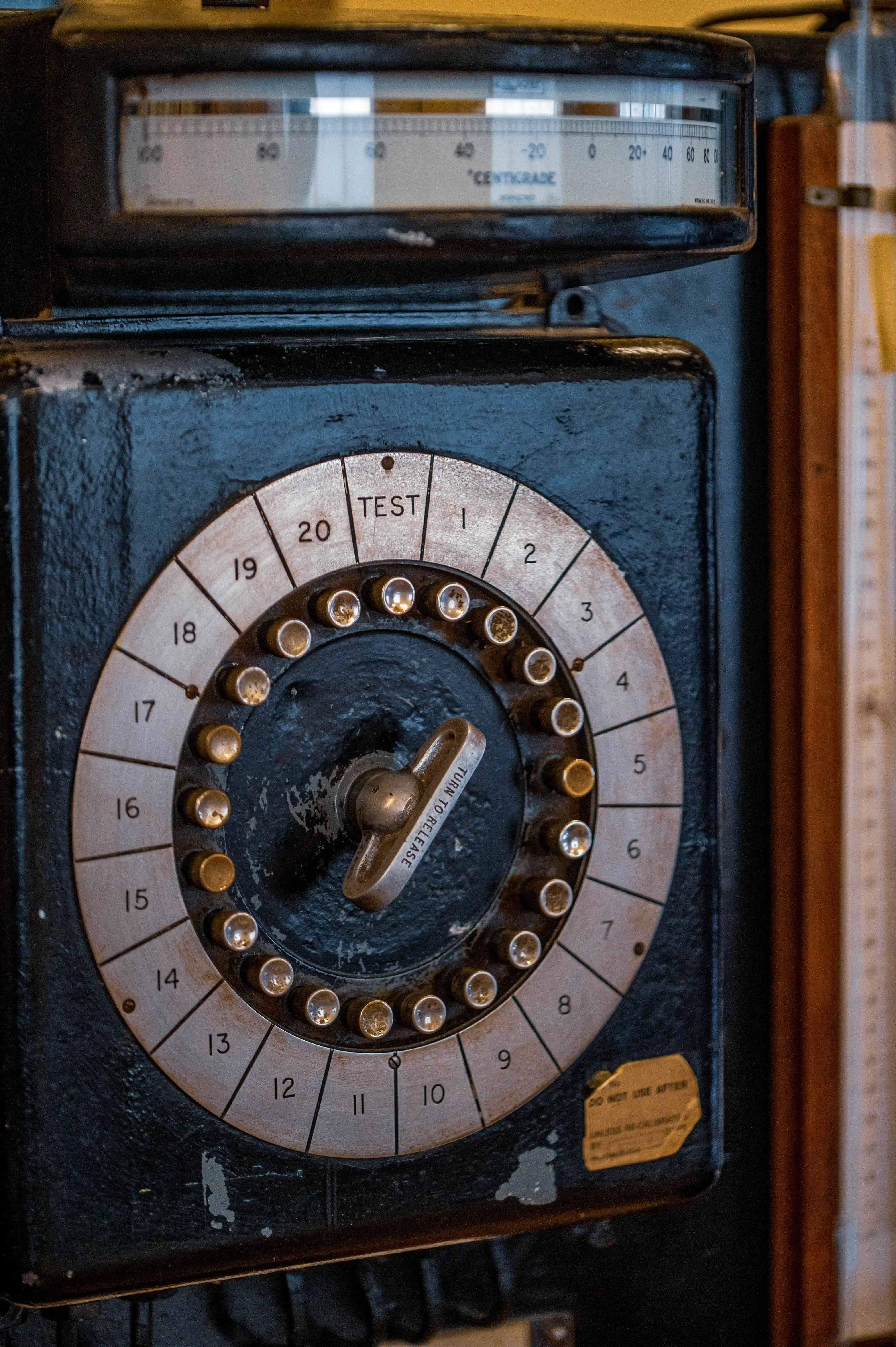 Close-up of a vintage black time clock. A circular dial with numbers 1-20, a TEST setting, and a metal handle labelled TURN TO RELEASE is central. Above, a thermometer scale reads CENTIGRADE from -20 to 100. A yellow caution sticker is on the lower right.