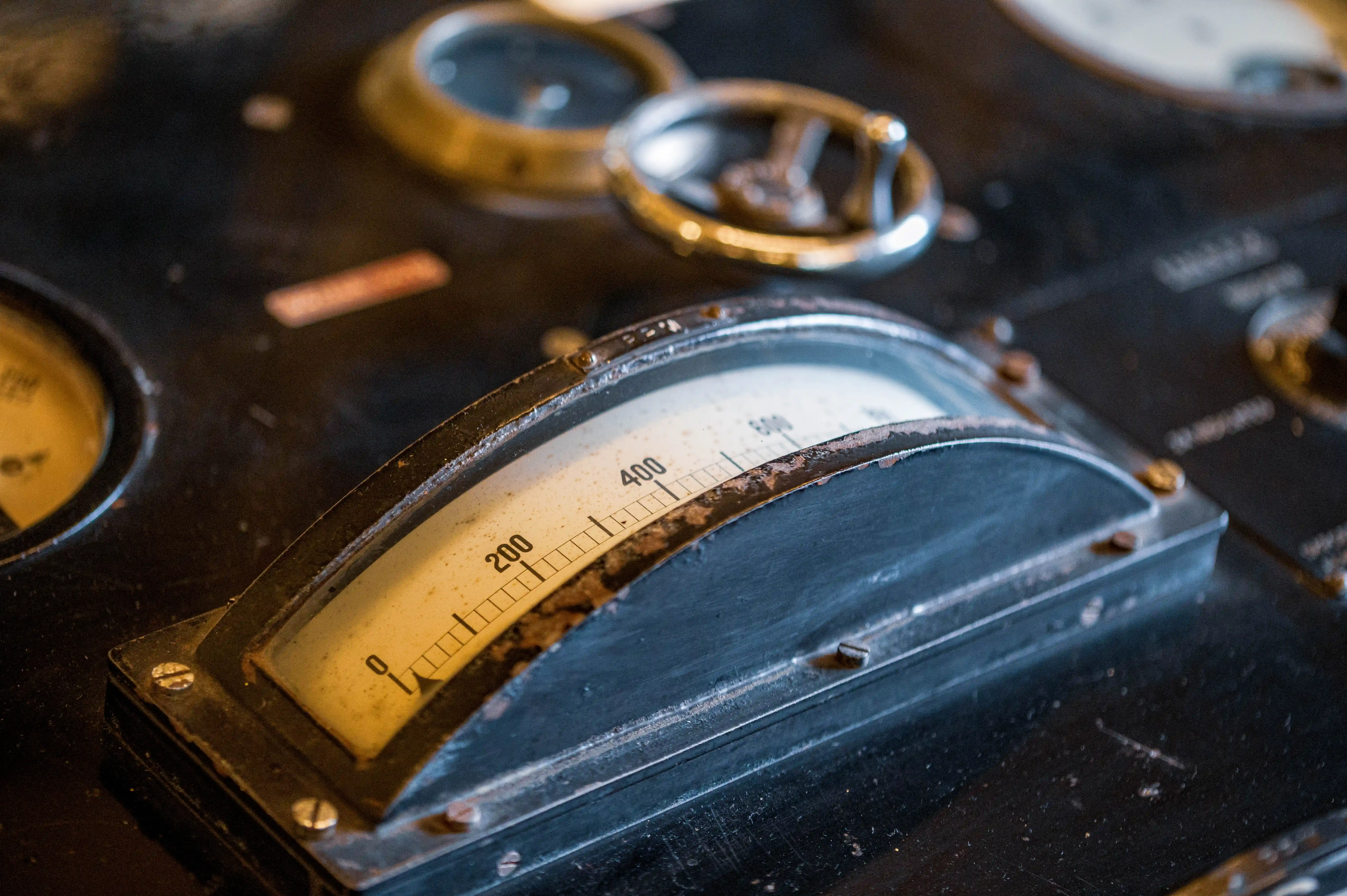 Close-up of a vintage analogue meter with a white face and black markings, showing a scale from 0 to 600. The meter is mounted on a dark, weathered control panel.