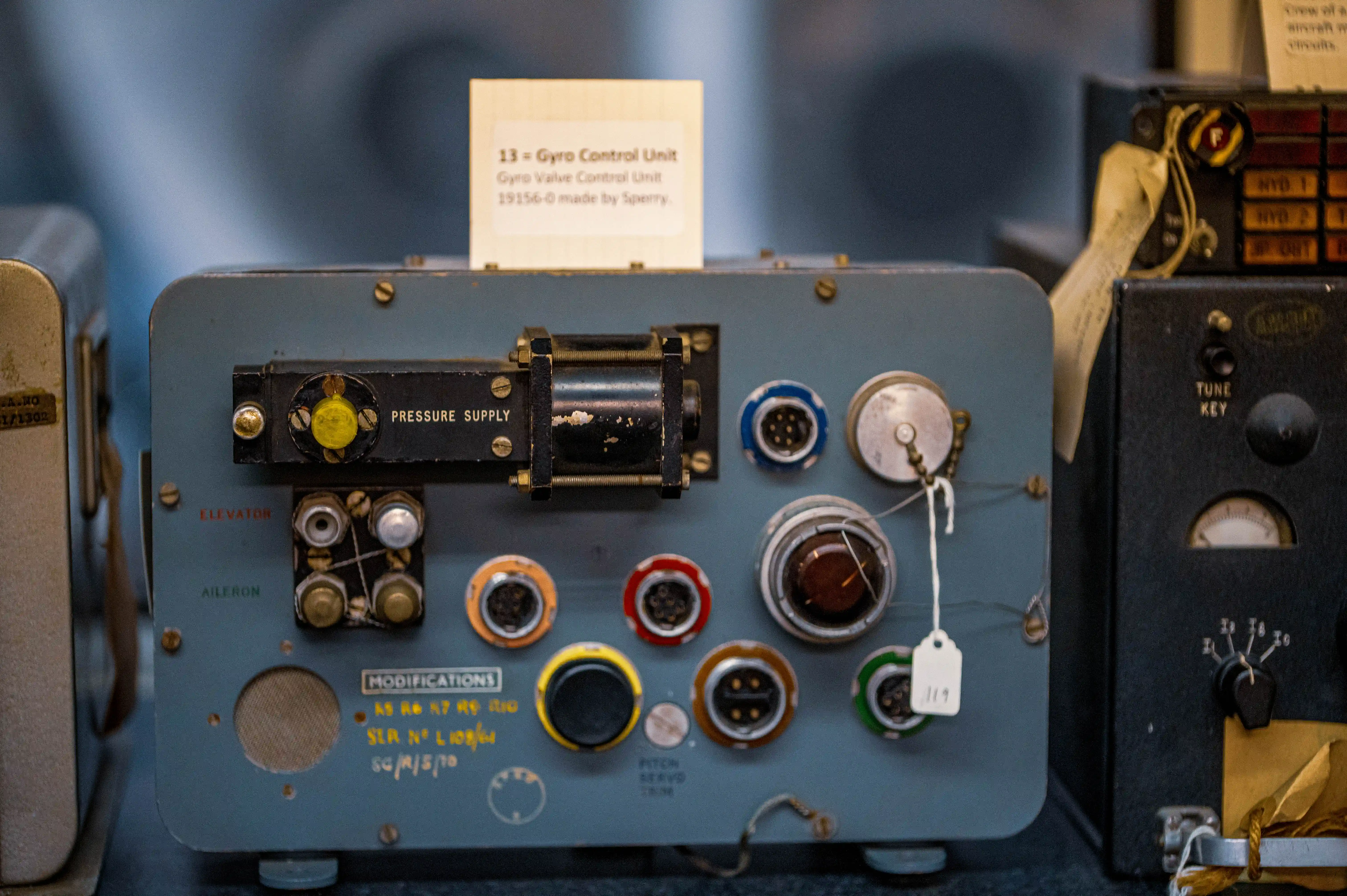 A vintage grey aircraft gyro control unit with various connectors, dials, and labels, including PRESSURE SUPPLY, ELEVATOR, AILERON, and PITCH SERVO. A card identifies it as a Gyro Control Unit made by Sperry.