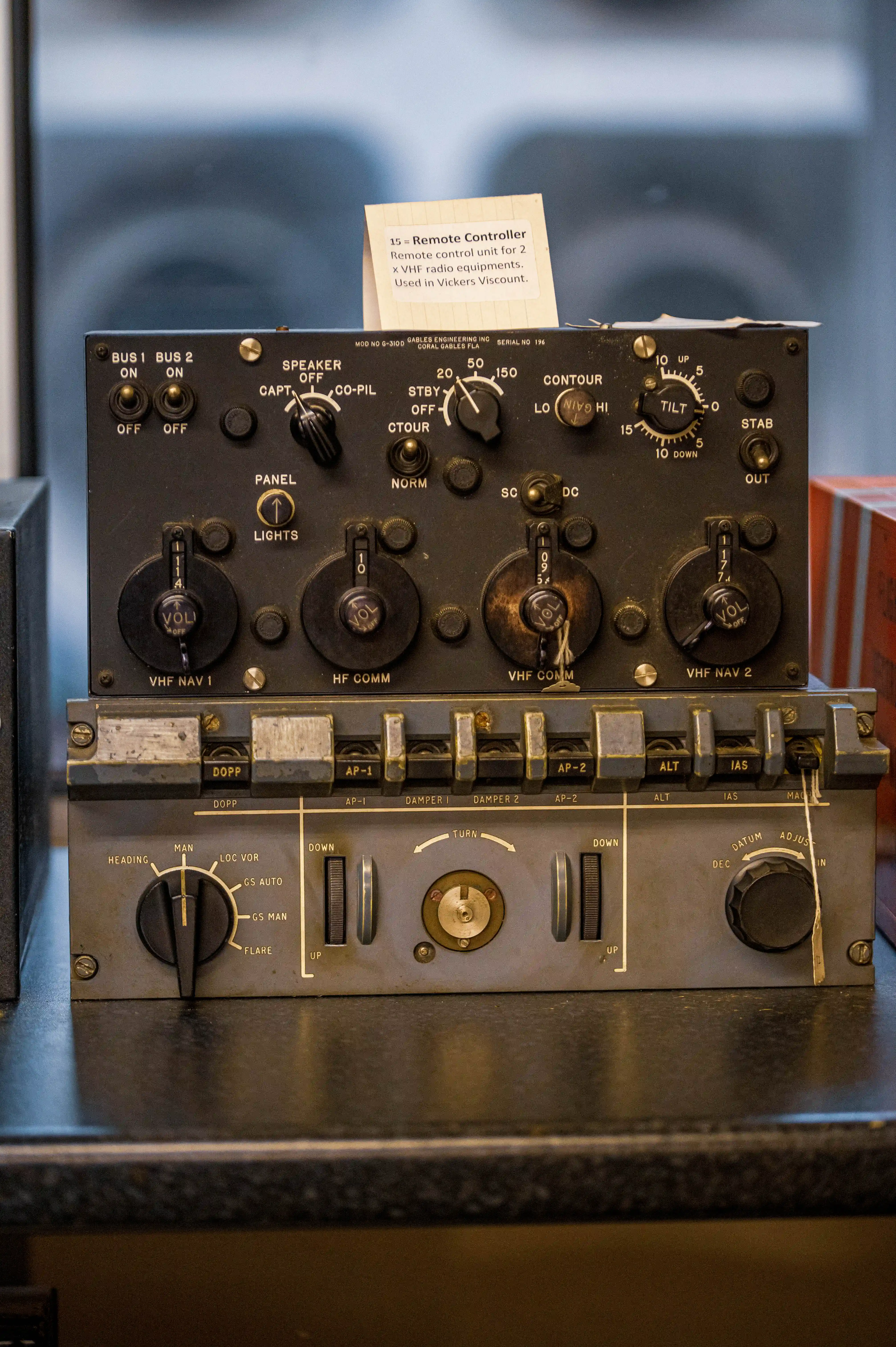 A detailed view of an old aircraft radio control panel. The panel is dark grey and covered in various controls, including rotary dials with numbers and labels like &laquo;VHF NAV 1,&raquo; &laquo;HF COMM,&raquo; and &laquo;VHF NAV 2.&raquo; Toggle switches and push buttons are also visible, along with indicators such as &laquo;BUS 1,&raquo; &laquo;BUS 2,&raquo; &laquo;SPEAKER,&raquo; &laquo;STBY,&raquo; and &laquo;ONTOUR.&raquo; A handwritten yellow label is attached to the top, stating its purpose and connection to a Vickers Viscount aircraft. The lower section of the image reveals another panel with more controls, including a large rotary dial labelled &laquo;HEADING&raquo; and smaller switches and dials.