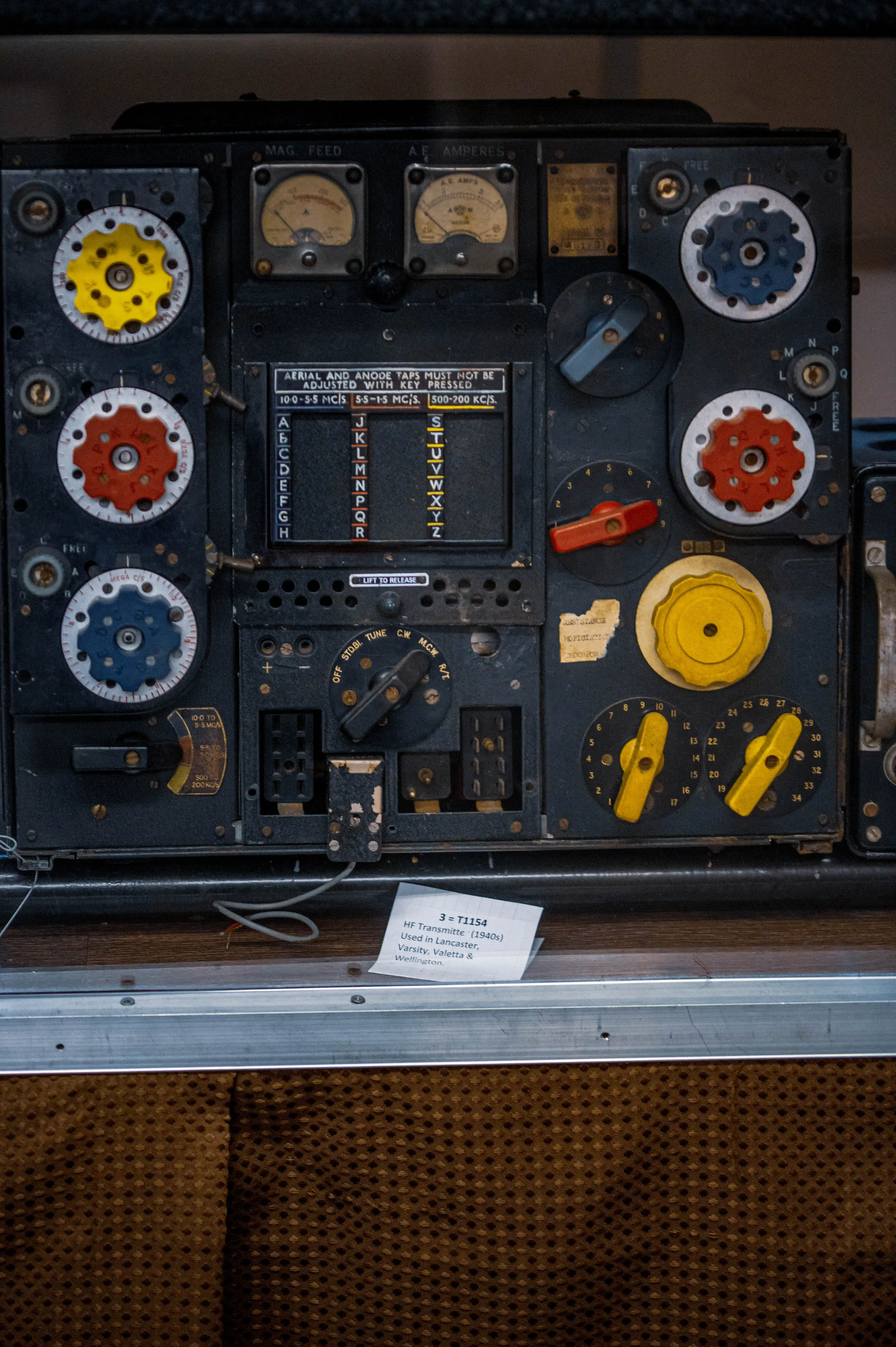A vintage aircraft radio control panel from the 1940s, featuring analog dials, switches, and knobs in a black metal housing. The panel includes color-coded controls (yellow, red, and blue), frequency meters, and labeled switches for tuning and adjusting radio settings. A handwritten note identifies the panel as a 'Type T1154 HF Transmitter' used in aircraft like the Lancaster, Varsity, Valetta, and Wellington.