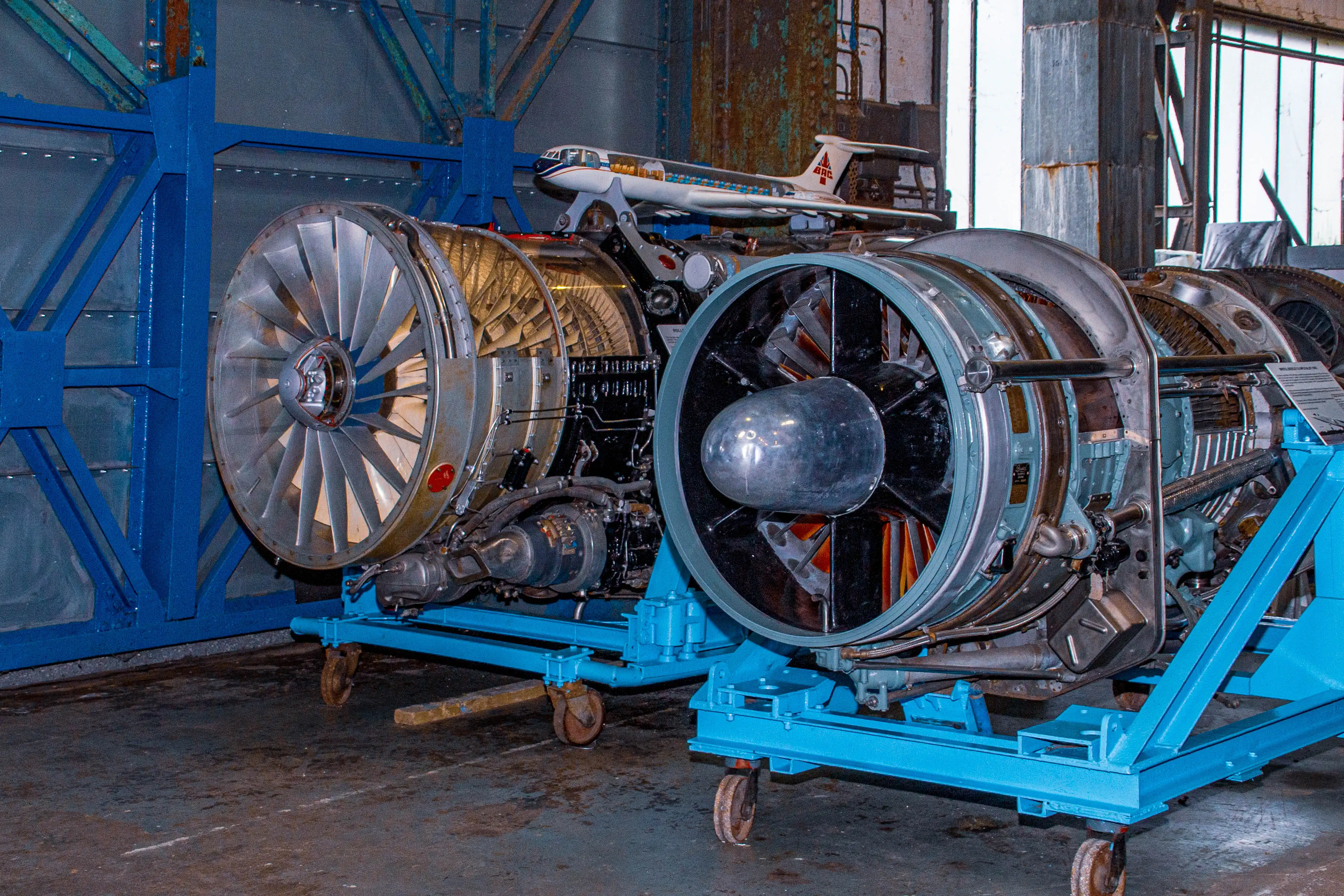 Two large, complex jet engines are side-by-side on blue trolleys. The engine on the left has a wide fan with many blades. The engine on the right has a darker, more rounded intake. A model aircraft sits above them.