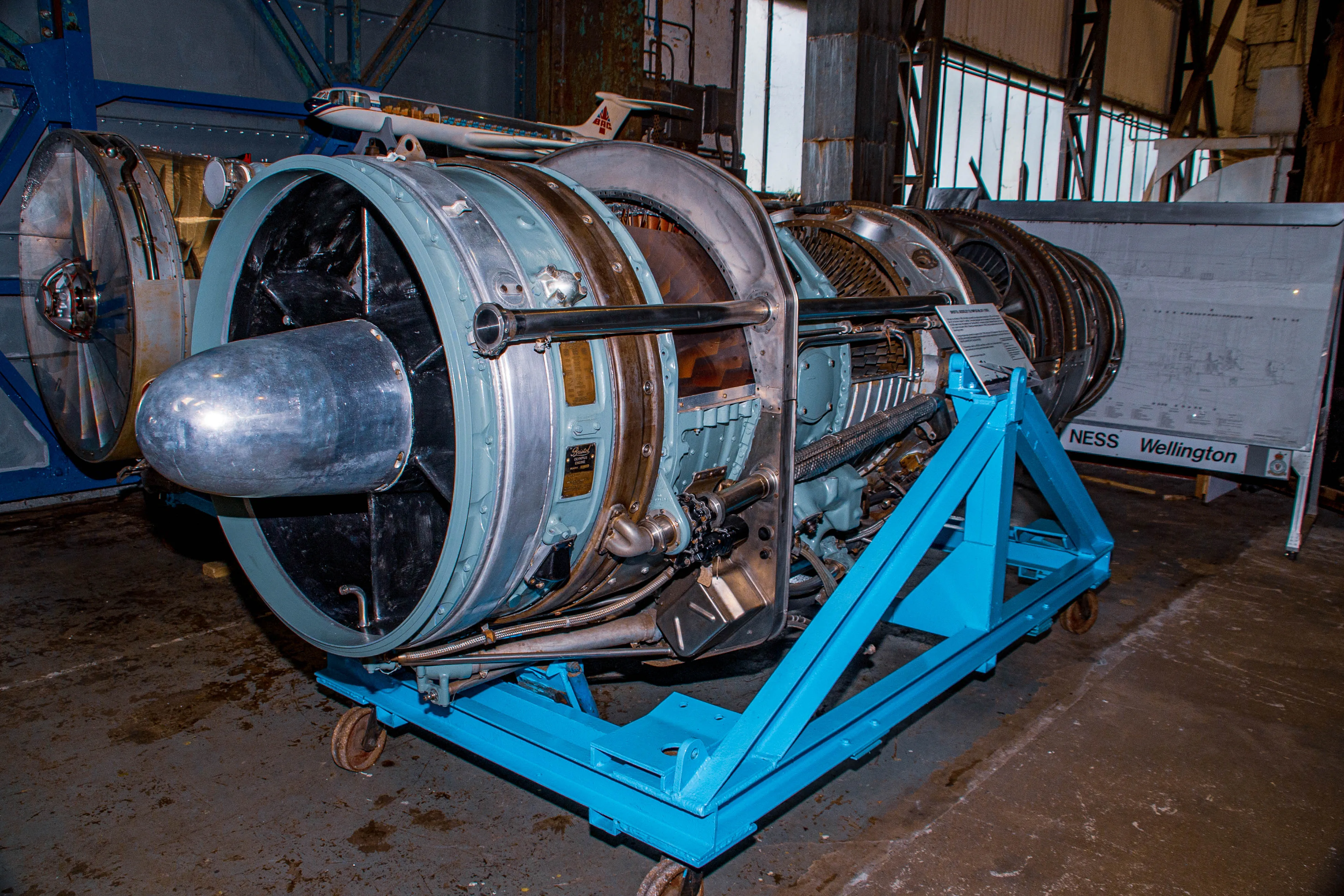 Close-up of a complex jet engine on a blue display stand, showing intricate details of its internal components and external piping. In the background, a model aeroplane and technical drawings are visible.