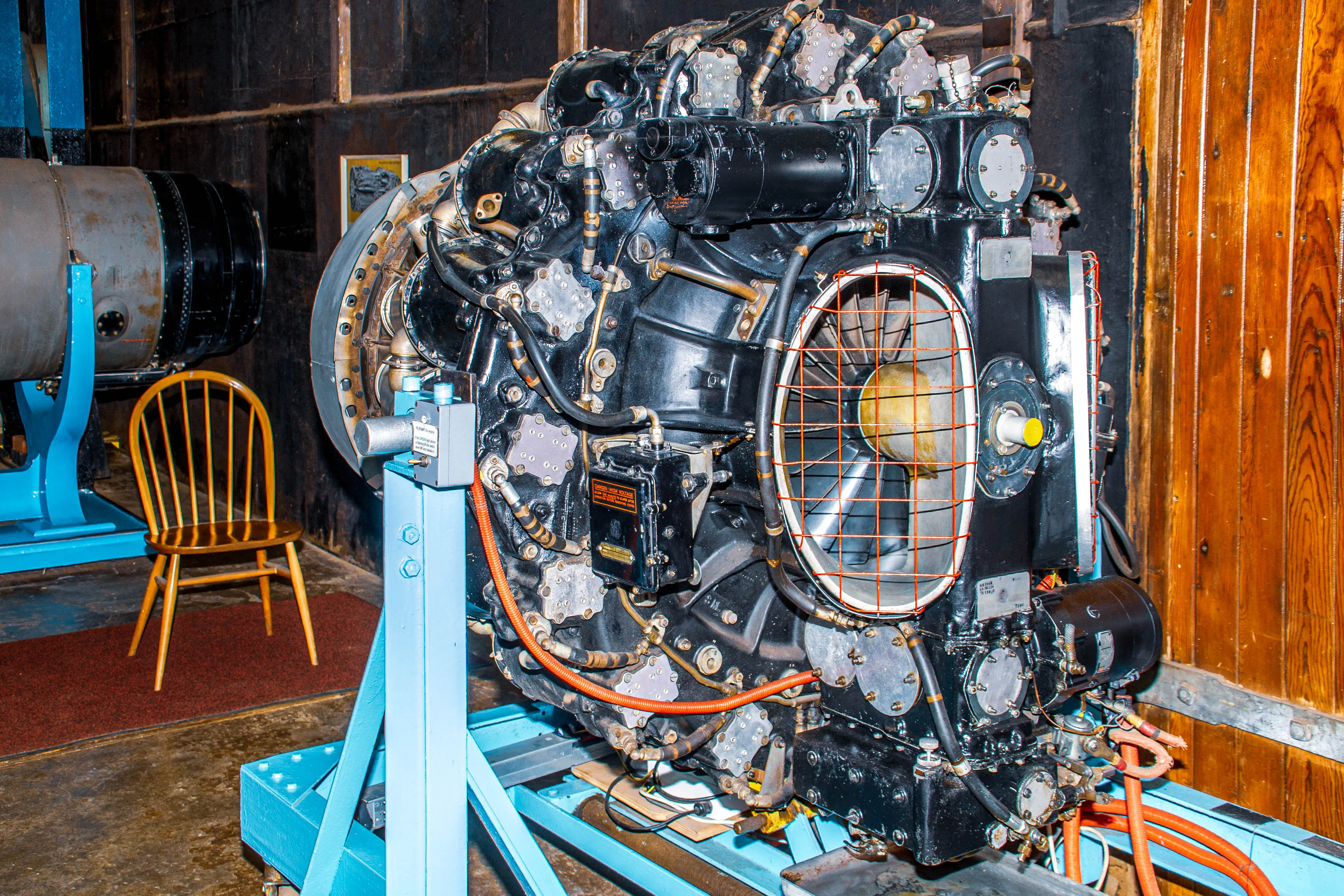 A close-up of a black radial aircraft engine with visible cylinders, pipes, wires, and a fan behind a wire mesh guard, all mounted on a blue support stand.