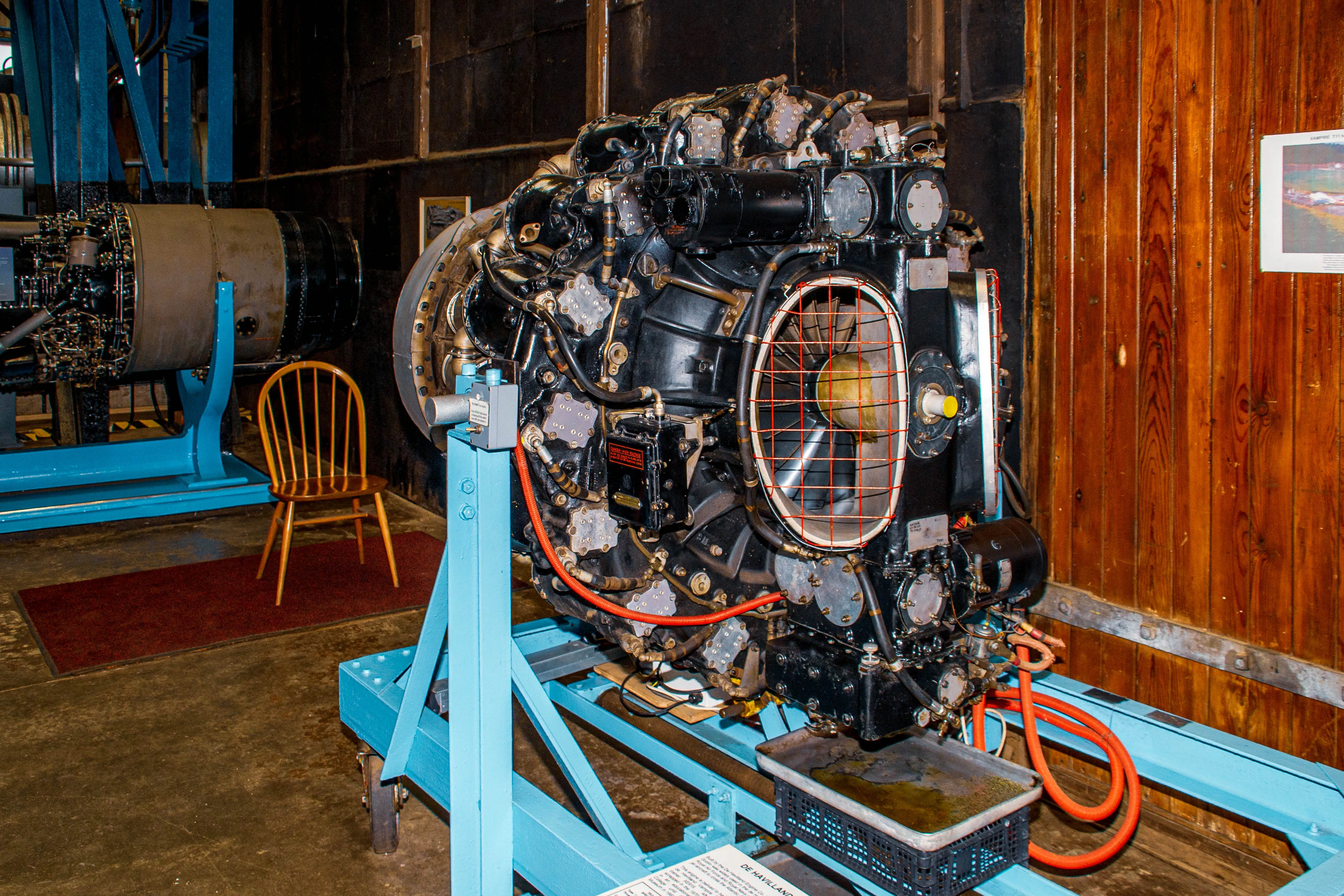 A close-up of a black radial aircraft engine with visible cylinders, pipes, wires, and a fan behind a wire mesh guard, all mounted on a blue support stand.