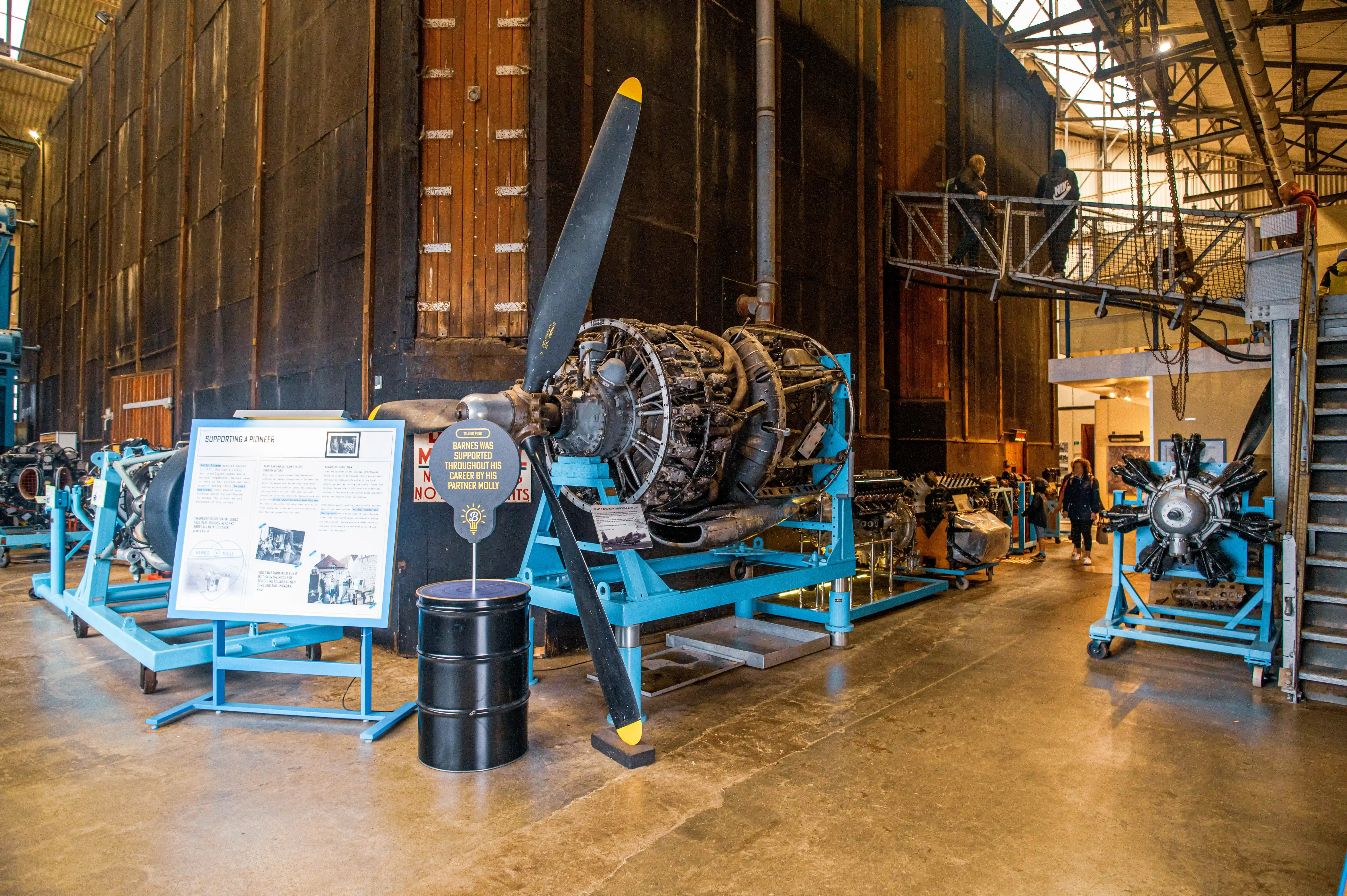A detailed view of a radial aircraft engine with its propeller, displayed indoors in a museum setting. An information placard stands next to the engine, and other exhibits are visible in the background.