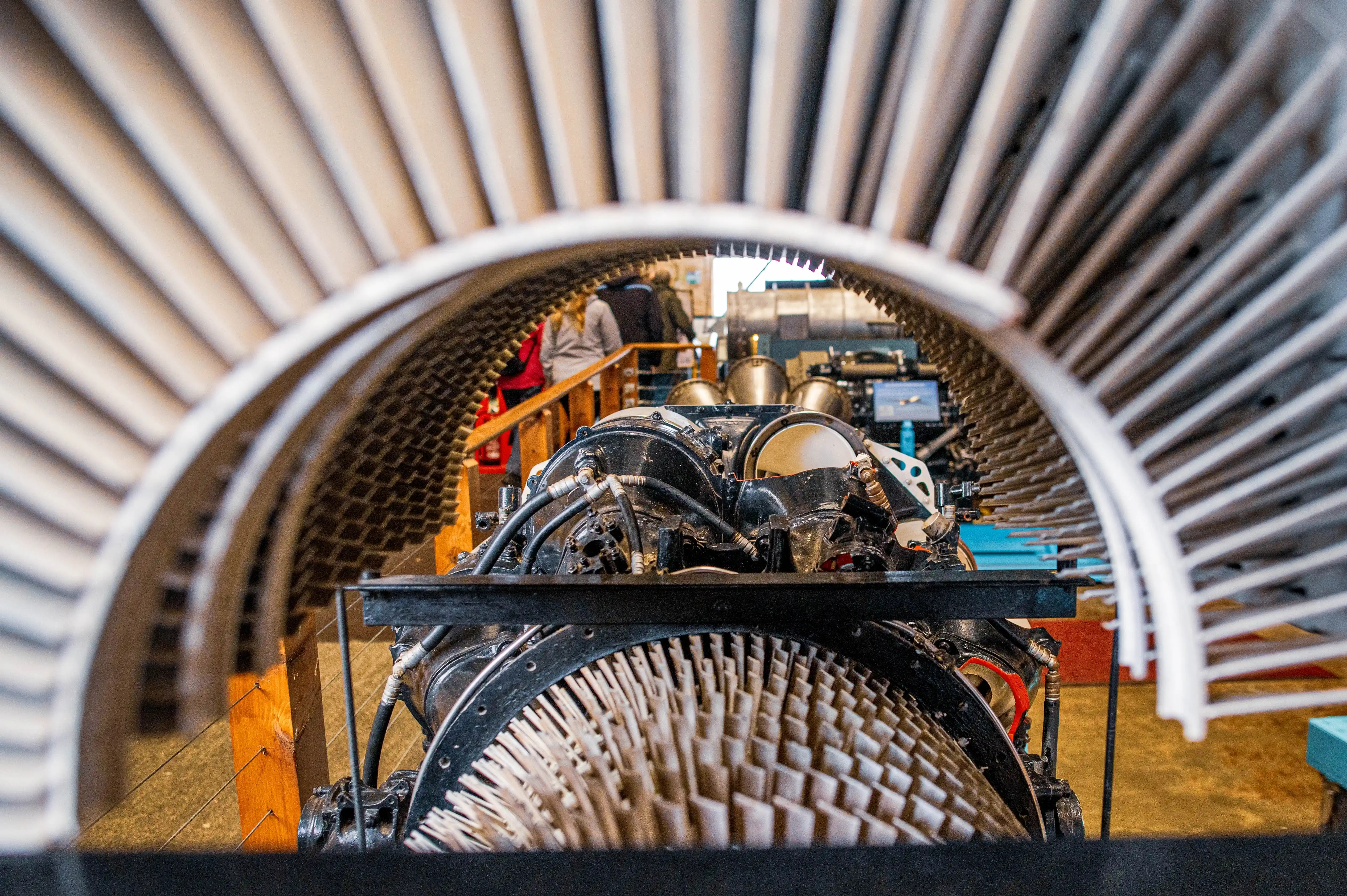 Close-up view of a jet engine's compressor blades, seen through the fan casing. Some visitors are visible in the blurred background.