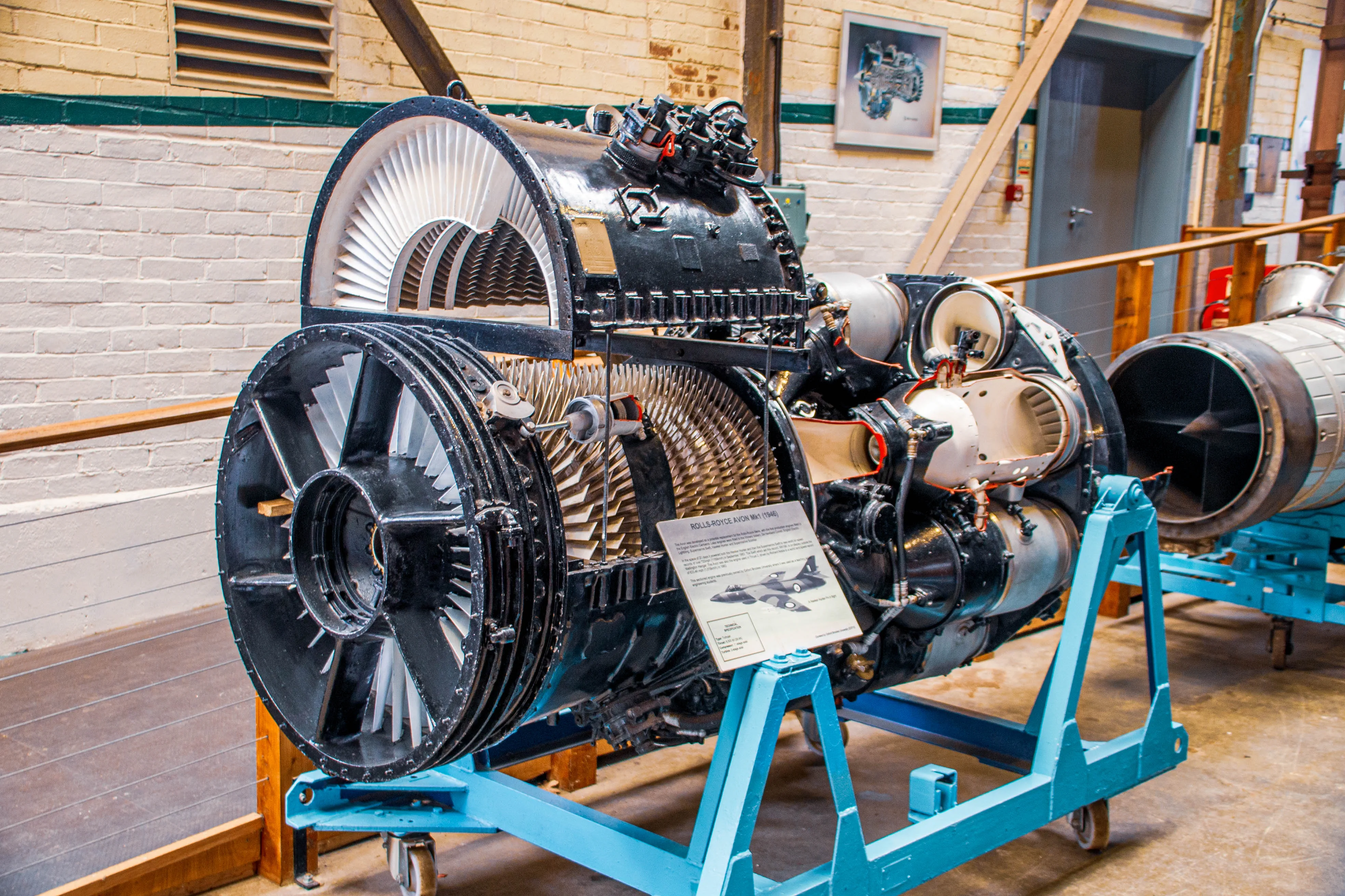 A detailed view of a Rolls-Royce Avon jet engine, mounted on a light blue stand in what appears to be a museum or industrial setting. The engine features a large fan at the front with a blue spinner, complex pipework, and a descriptive plaque.