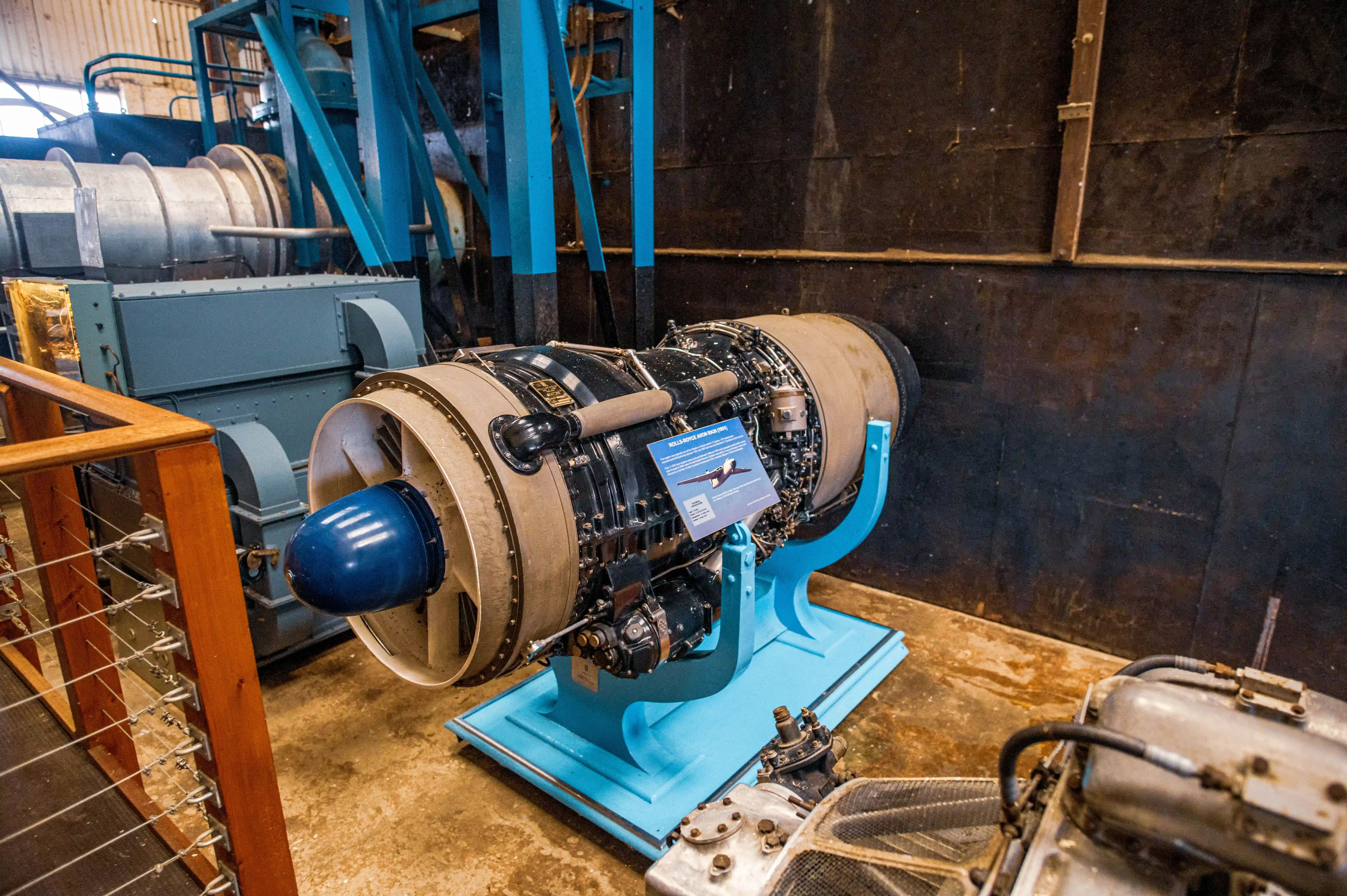 A detailed view of a Rolls-Royce Avon jet engine, mounted on a light blue stand in what appears to be a museum or industrial setting. The engine features a large fan at the front with a blue spinner, complex pipework, and a descriptive plaque.