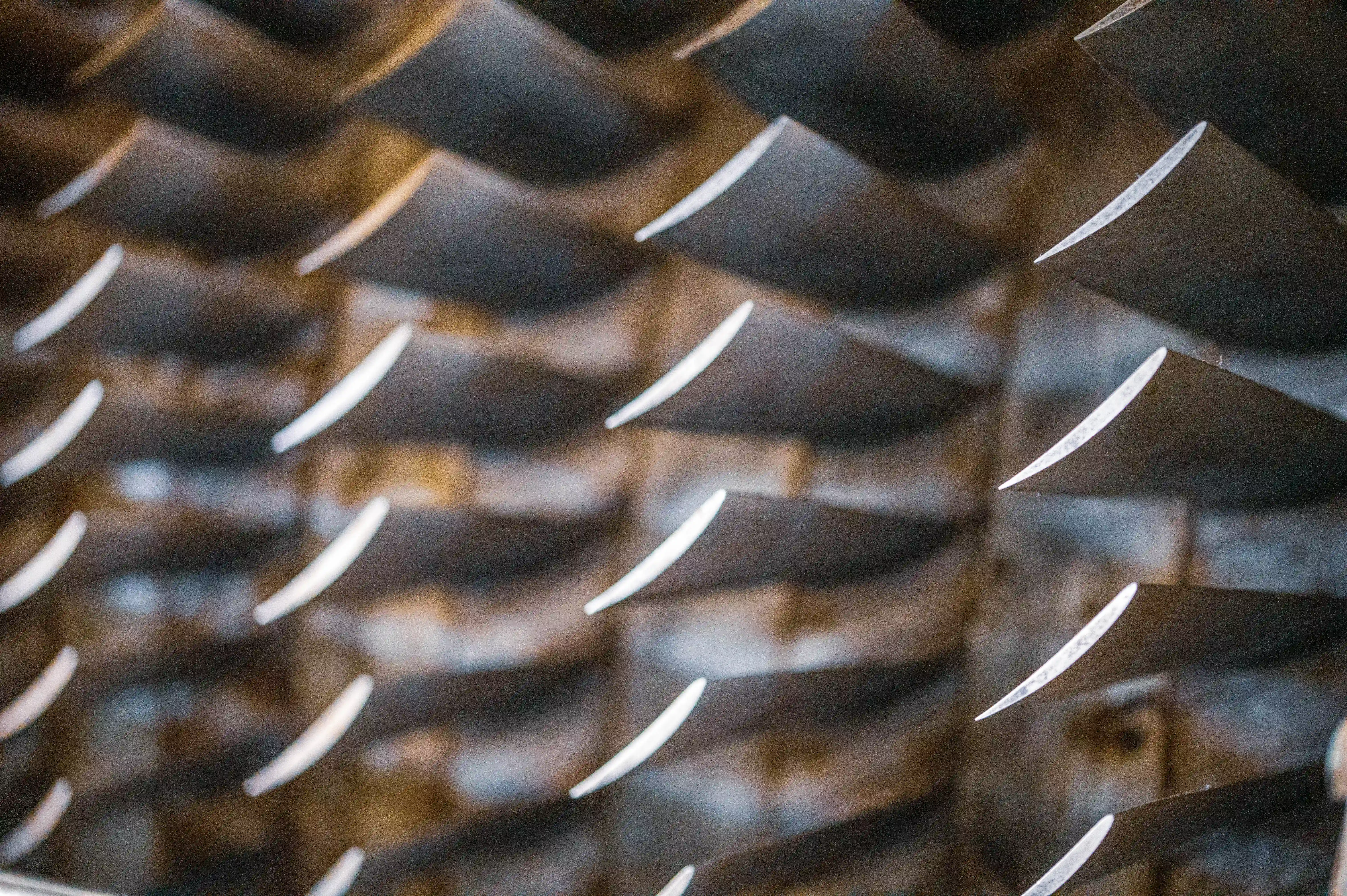 Close-up, abstract view of multiple metallic turbine blades arranged in a grid pattern. The blades have a polished, slightly reflective surface and are set against a darker, textured background.
