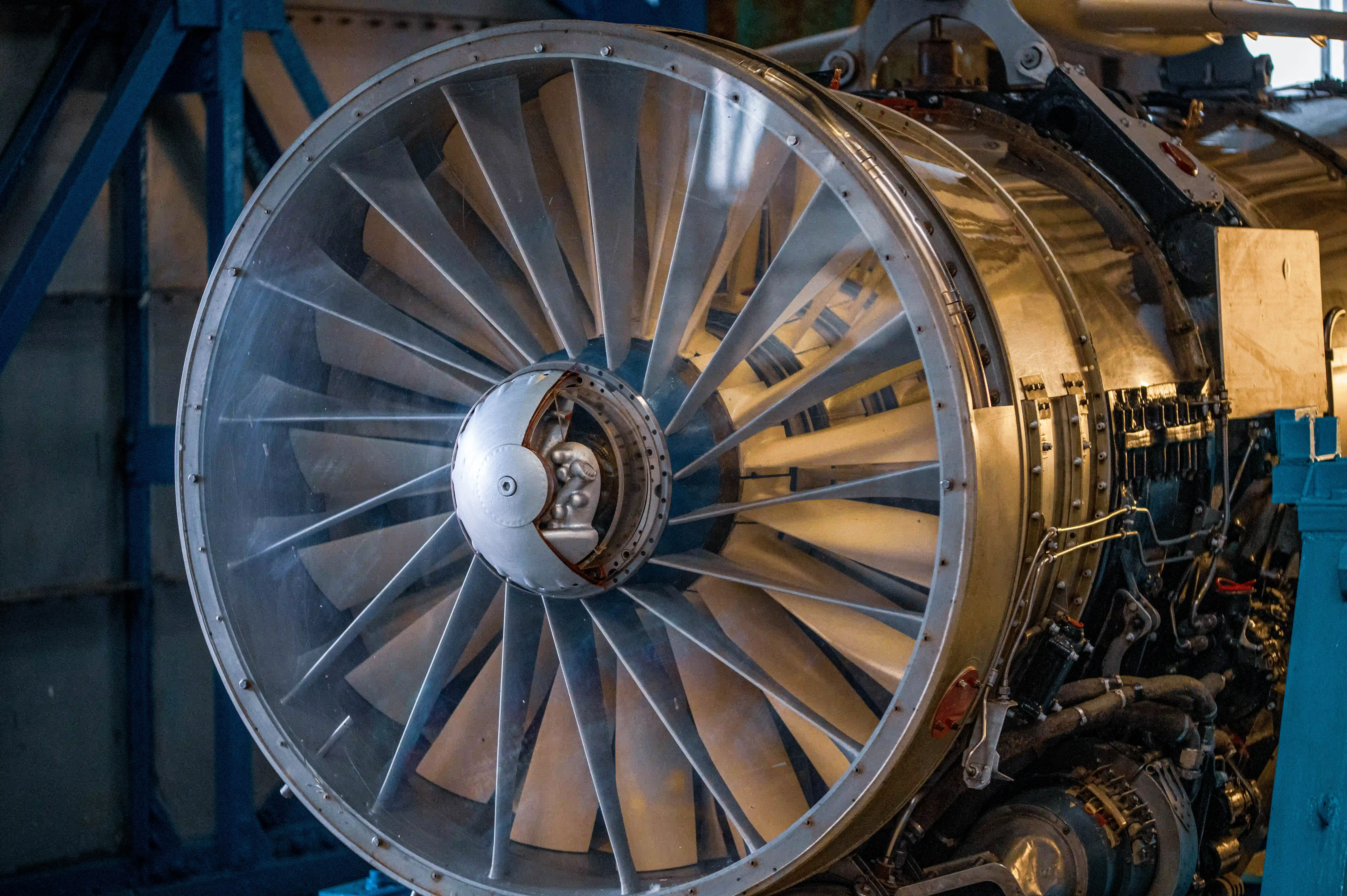 Close-up of the front fan of a jet engine, with multiple polished metal blades fanning out from a central hub. The engine's complex internal components and fuel lines are visible behind the fan blades. The engine is situated within a blue industrial support structure.