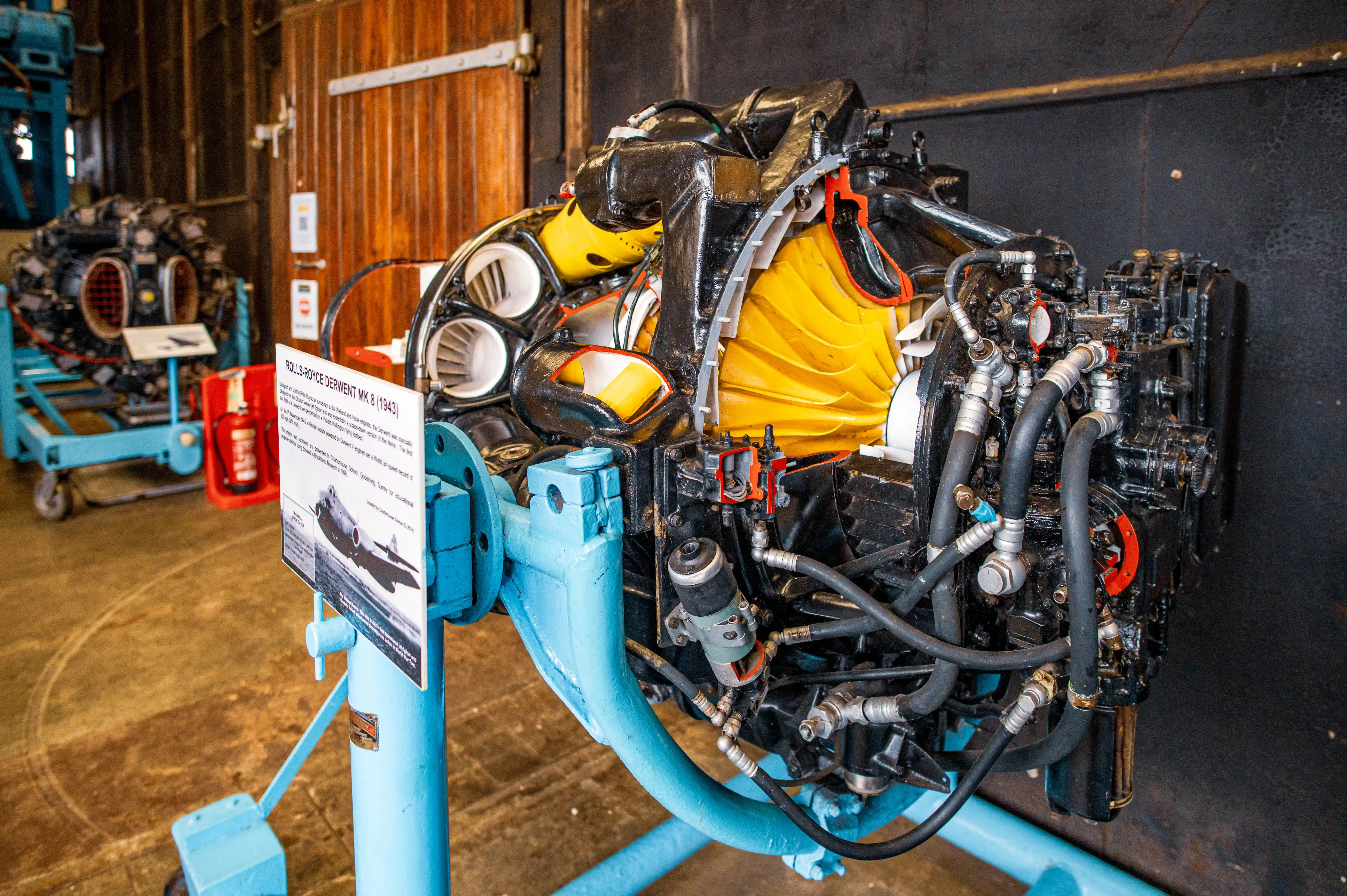 A detailed cutaway model of a black and yellow Rolls-Royce Derwent Mk 8 jet engine, mounted on a bright blue stand, in an exhibition setting.