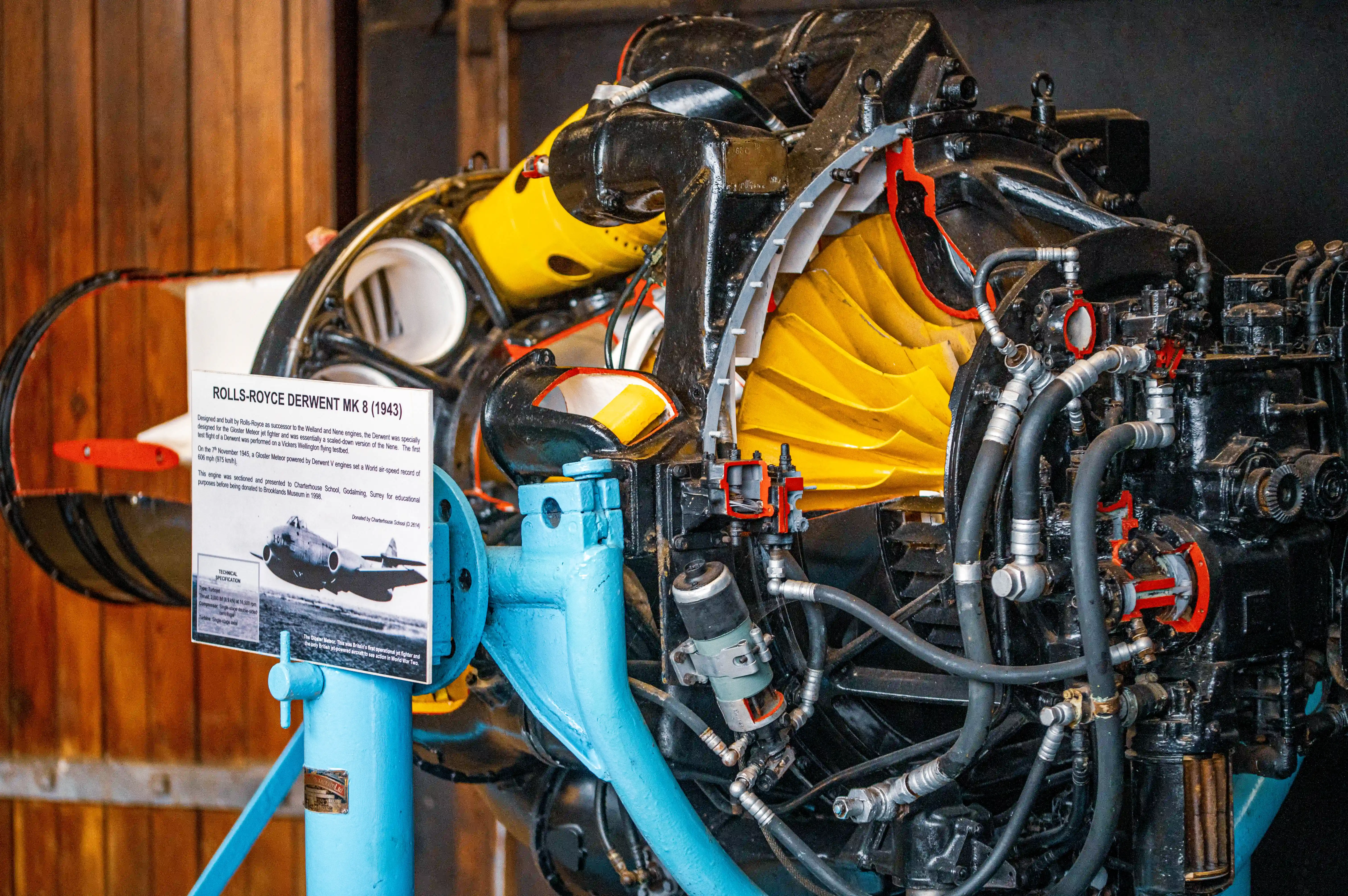 A detailed cutaway model of a black and yellow Rolls-Royce Derwent Mk 8 jet engine, mounted on a bright blue stand, in an exhibition setting.