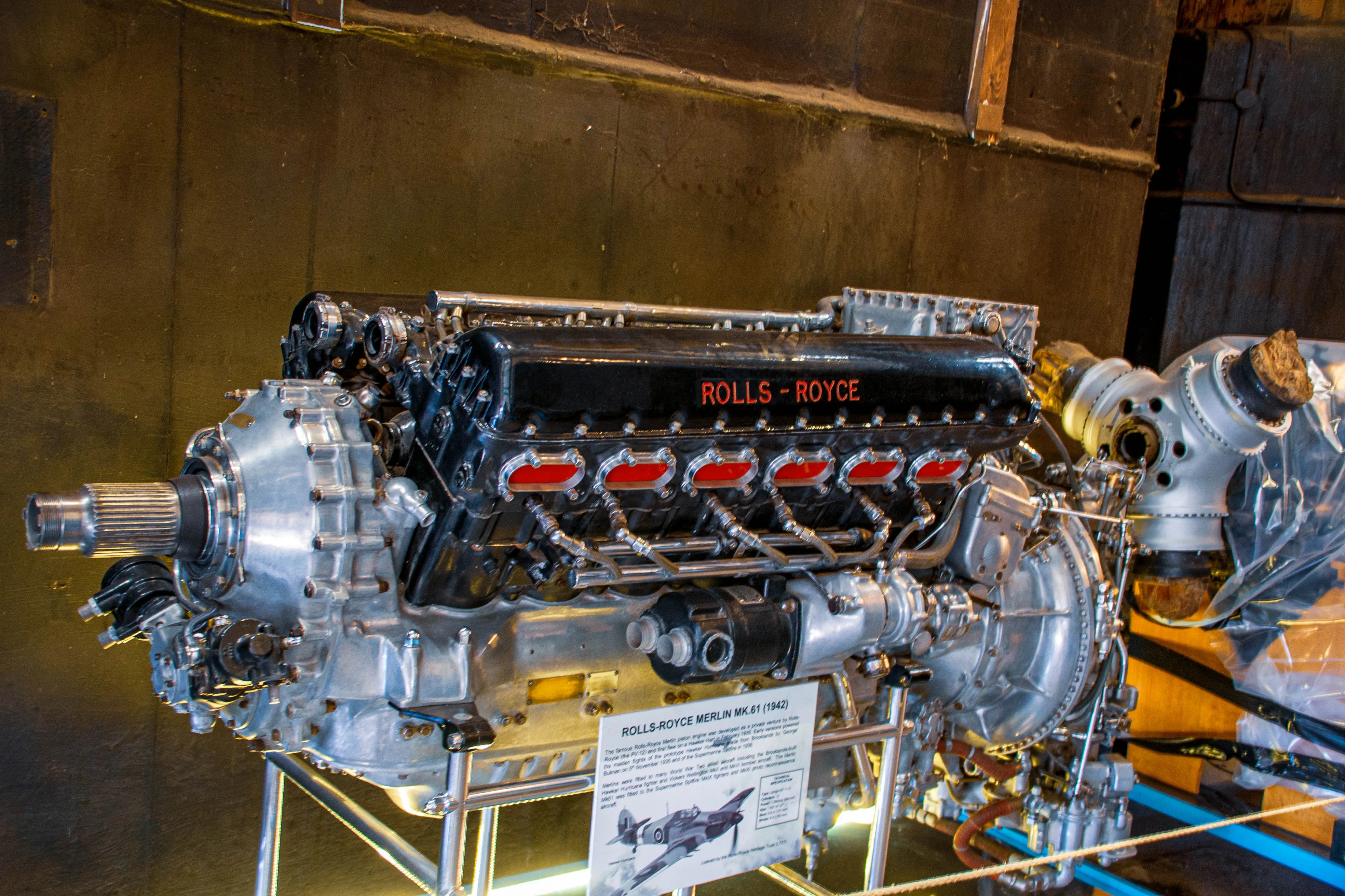 A Rolls-Royce Merlin MK.61 engine (1942) displayed in a museum setting, showcasing its iconic V12 design with red valve covers and polished metal components. The engine is mounted on a metal stand with an informational plaque detailing its historical significance. The background features a dark wooden wall.
