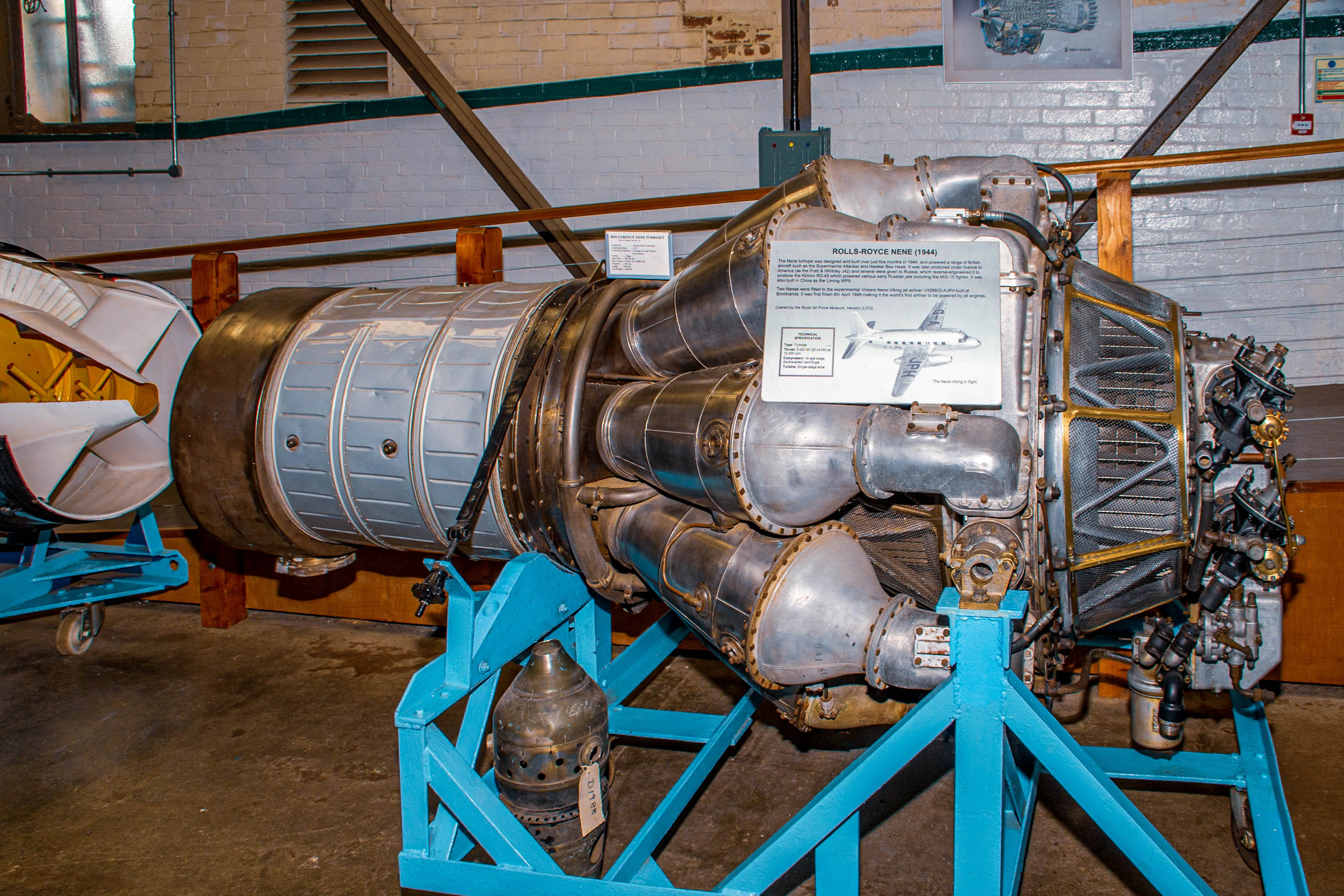 A detailed cutaway model of a black and yellow Rolls-Royce Derwent Mk 8 jet engine, mounted on a bright blue stand, in an exhibition setting.