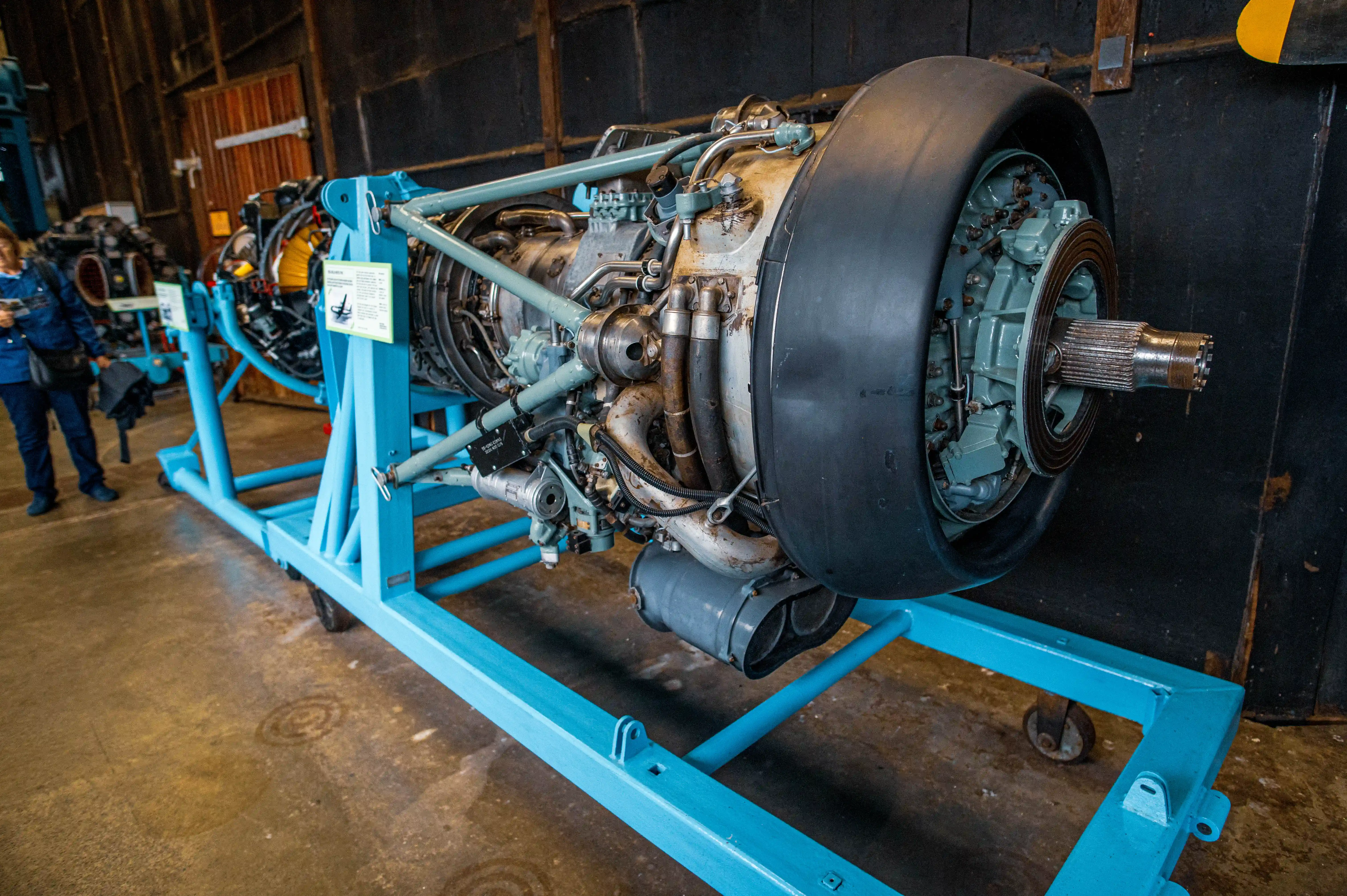A detailed cutaway model of a black and yellow Rolls-Royce Derwent Mk 8 jet engine, mounted on a bright blue stand, in an exhibition setting.