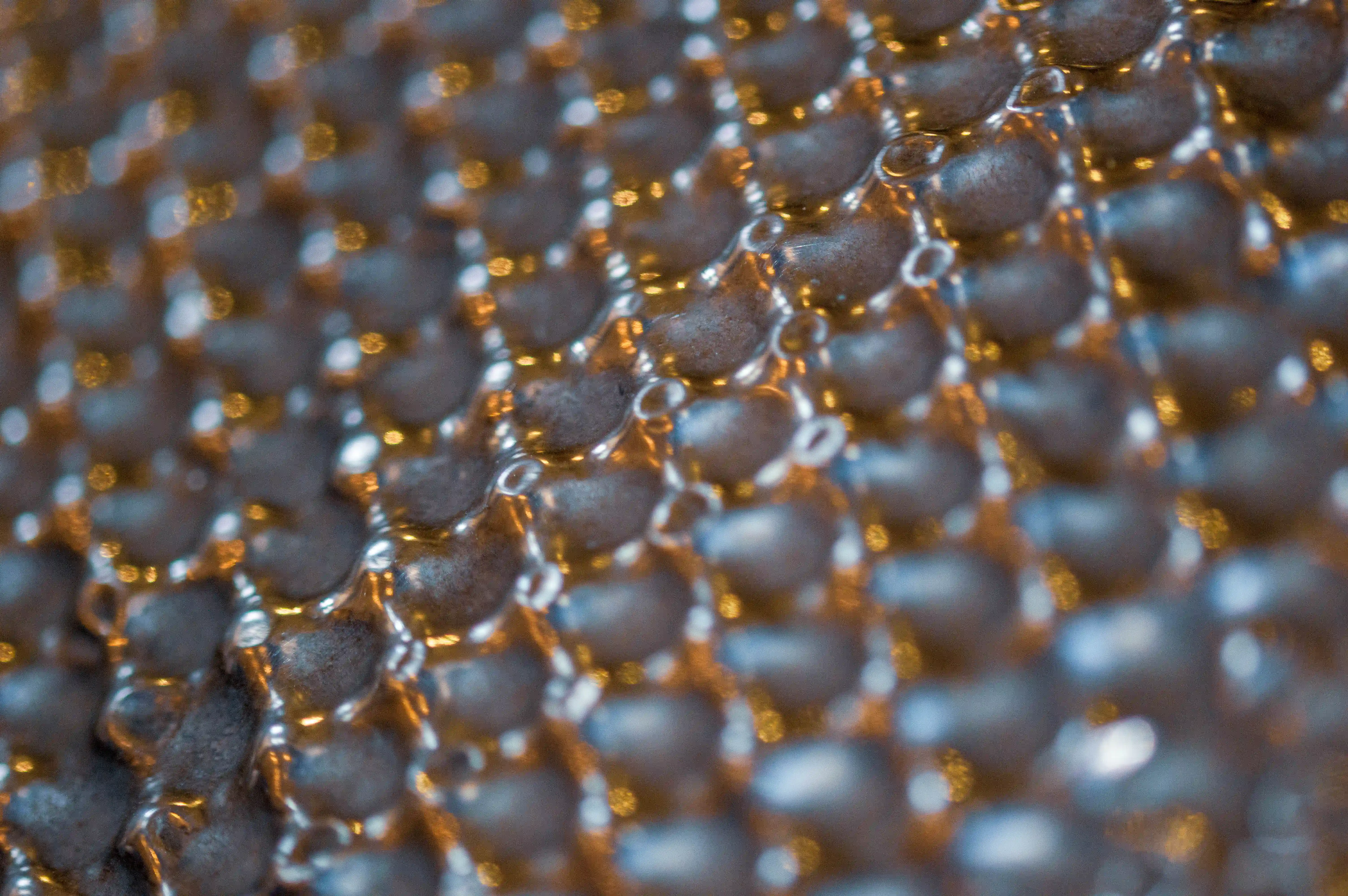 Extreme close-up shot of a dark, textured surface with a repeating hexagonal pattern. Small, rounded shapes, possibly links of a chainmail or a honeycomb structure, are visible. Bright, out-of-focus golden and white highlights are scattered across the surface, creating a bokeh effect and highlighting the three-dimensional quality of the material.