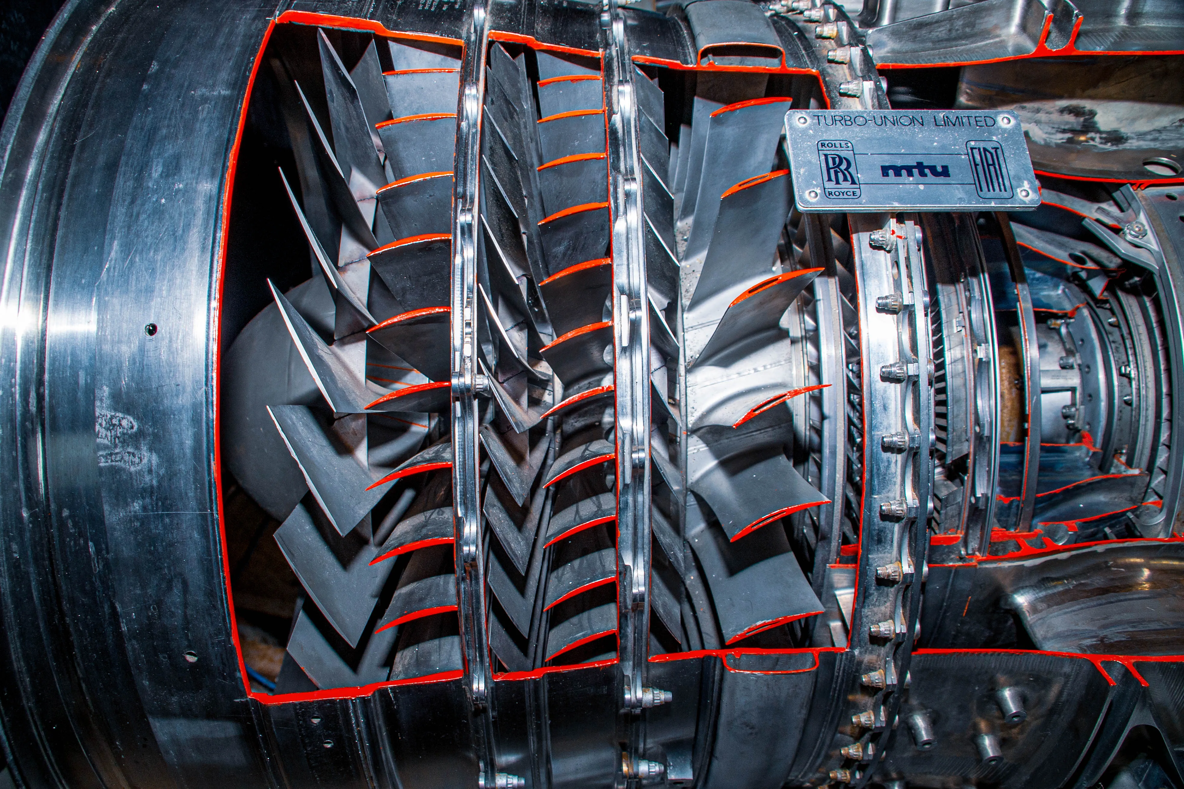 Close-up view of a jet engine's interior, showing multiple rows of grey fan blades with red edges. A metal plaque with 