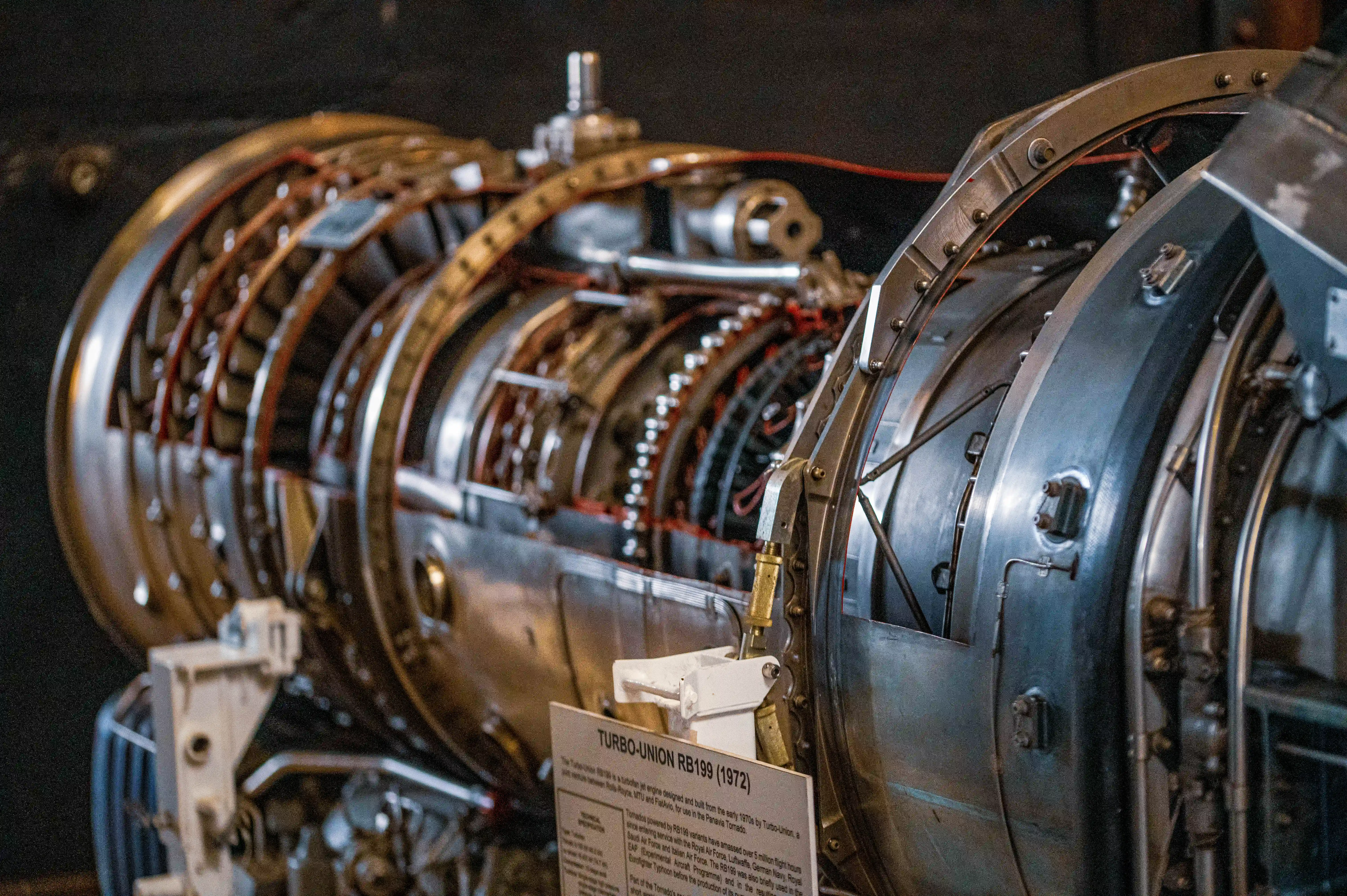 A close-up photograph showing the intricate internal workings of a Turbo-Union RB199 jet engine. Multiple stages of turbine and compressor blades are visible, along with various metal casings, pipes, and electrical wiring. A descriptive plaque is partially visible in the foreground.