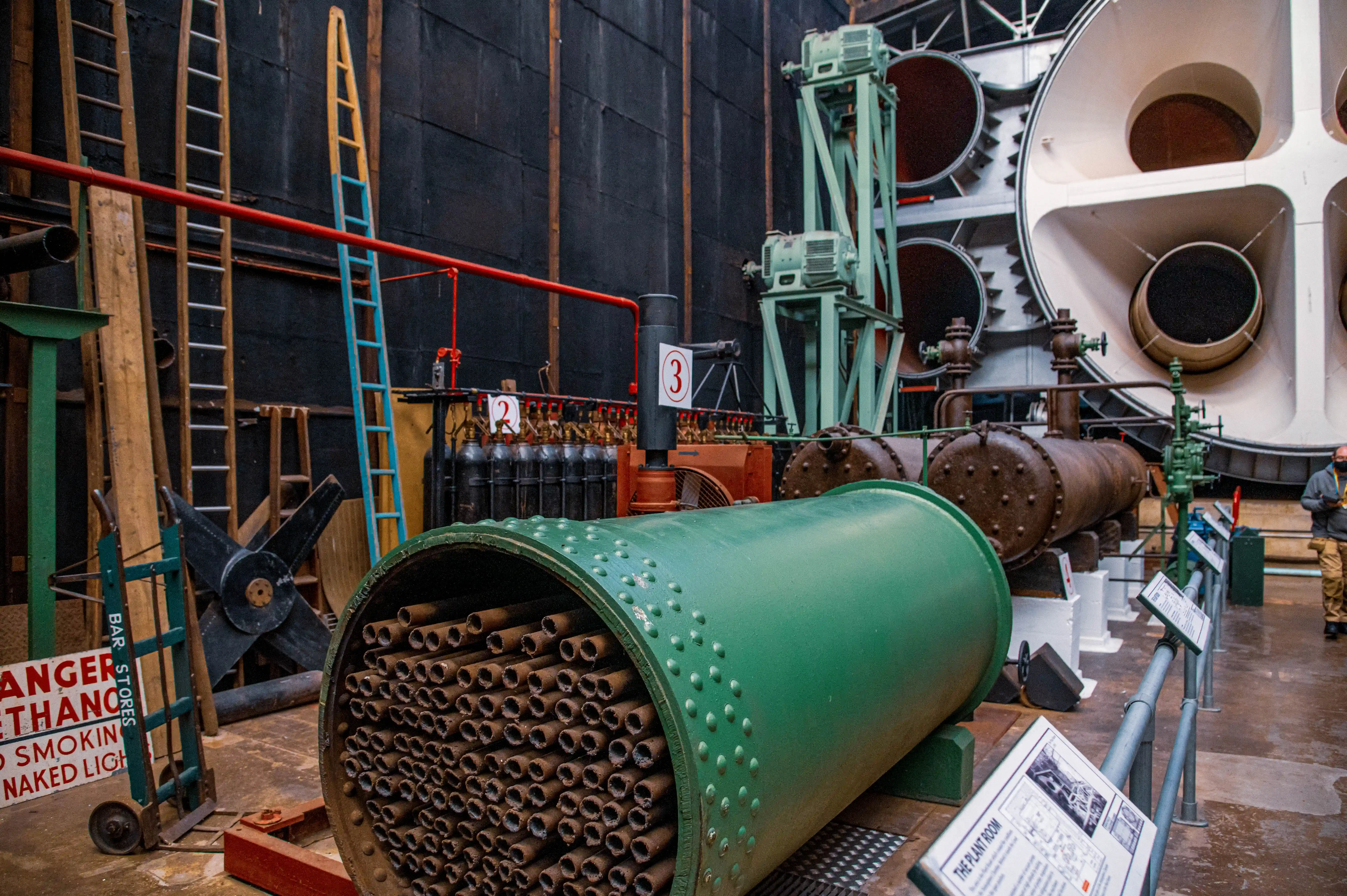 Industrial machinery displayed in a museum, including a green cylindrical unit with internal tubes, numbered tanks and surrounding pipes.