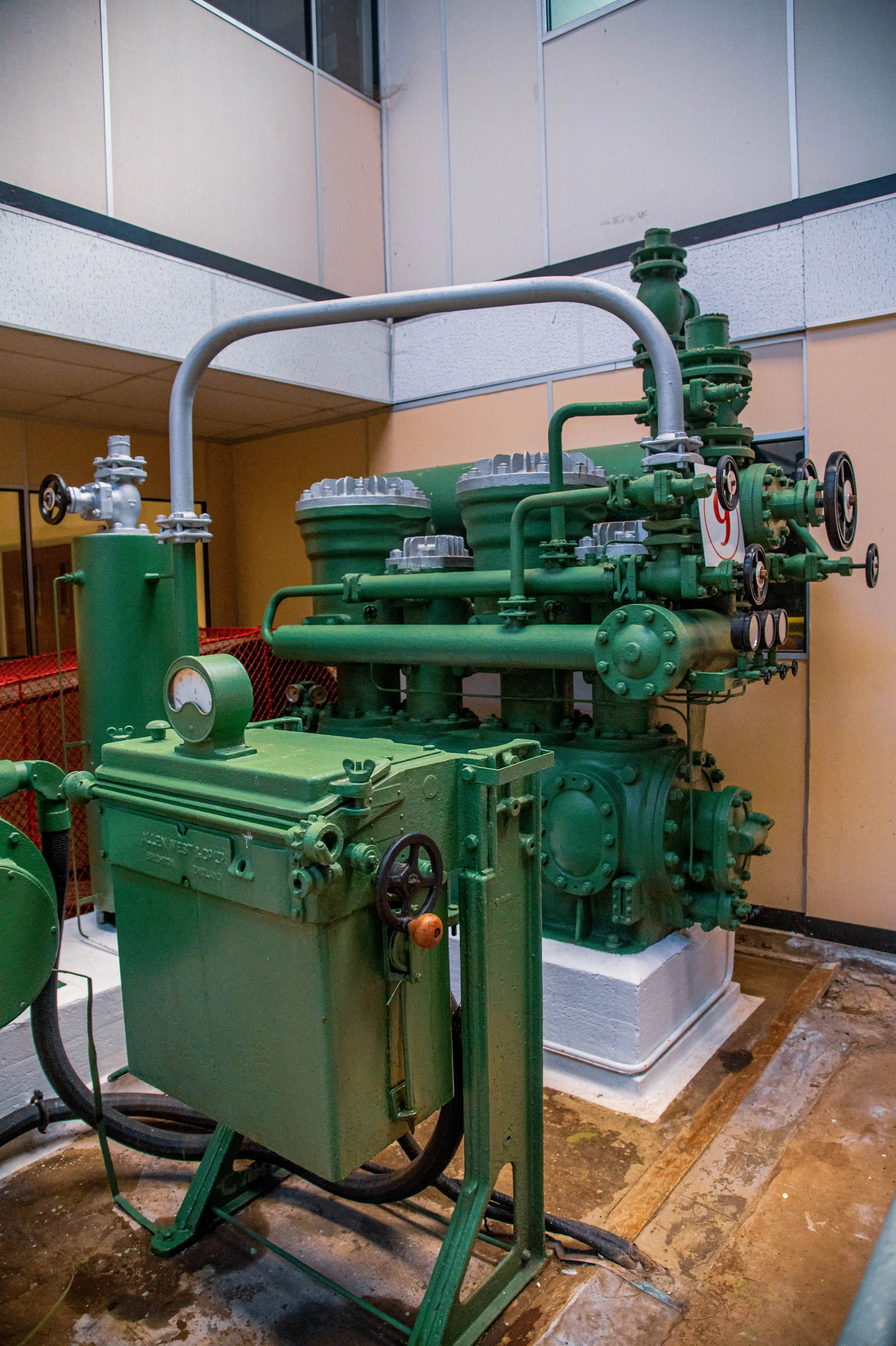 Green industrial compressor with pipes, valves and a pressure gauge displayed indoors behind a red safety barrier.