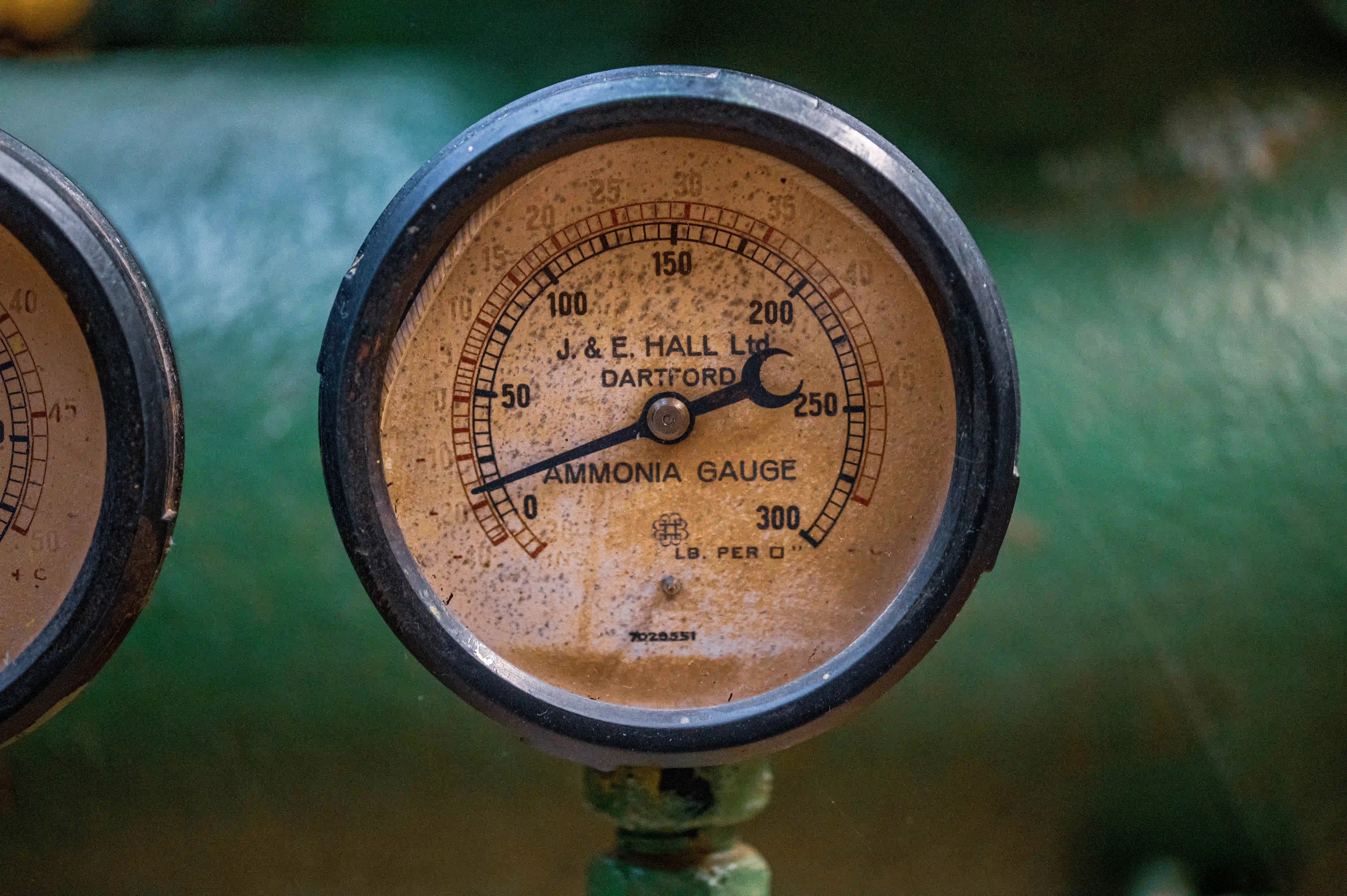 A close-up of an old ammonia pressure gauge with a dial face showing markings from 0 to 300 lb. per sq. in. and a needle indicating around 120 lb. per sq. in.