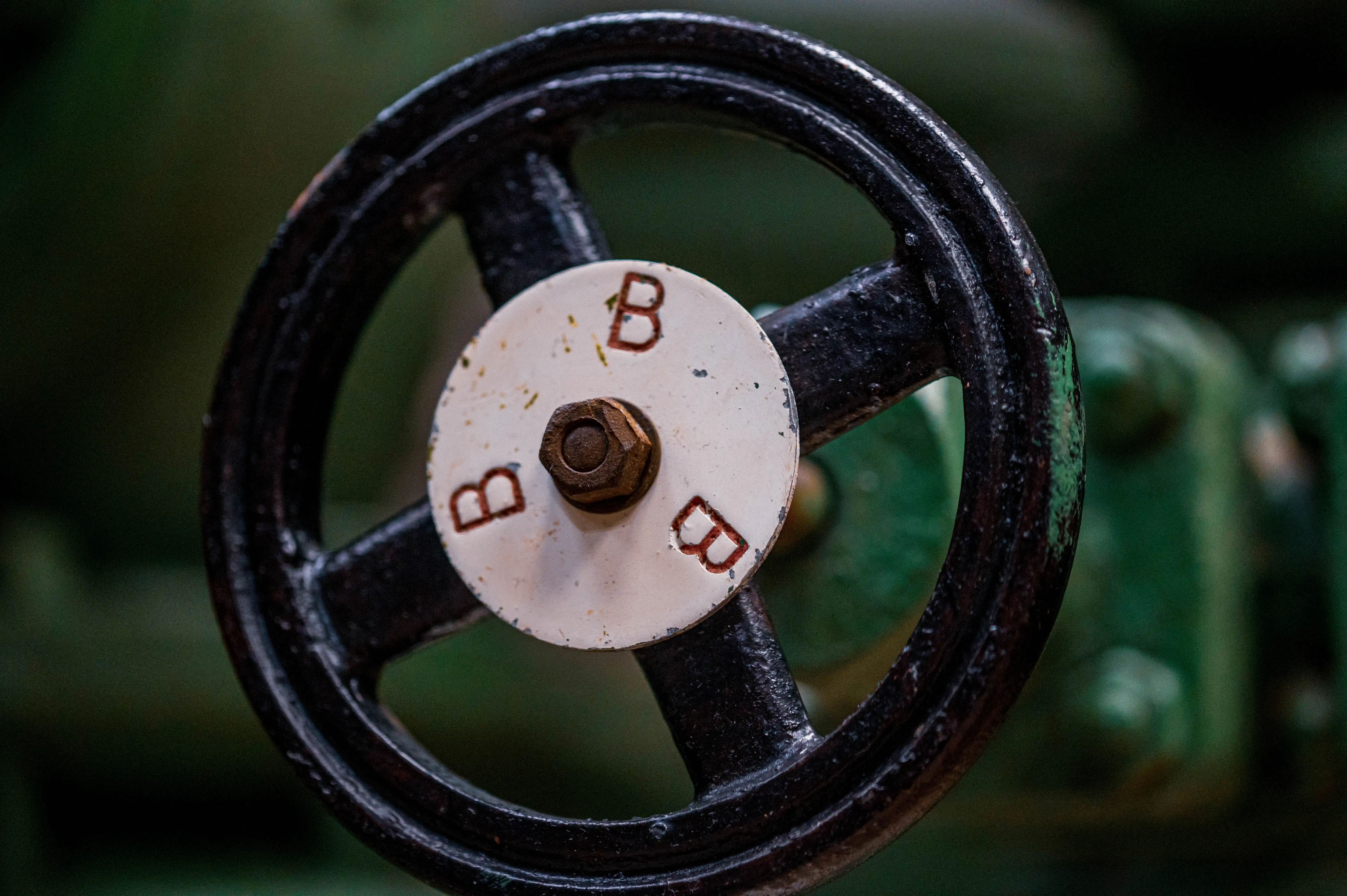 A close-up photograph of a black, industrial valve wheel with a white central cap. The cap features three red stencilled letters 'B' around a rusted bolt. The background is out of focus and a muted green colour.
