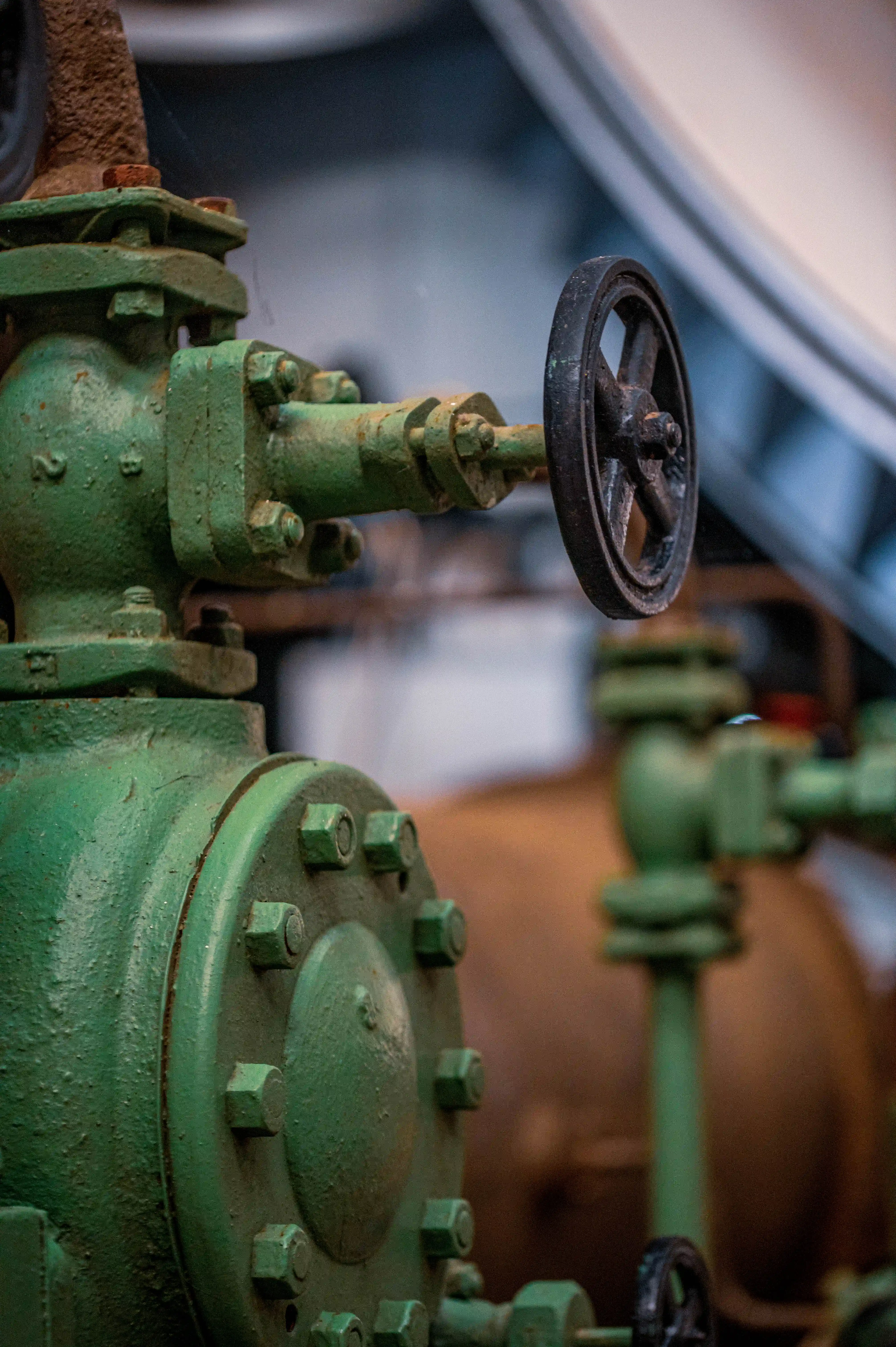 Close-up of a vintage green industrial valve with a black wheel, showing a textured surface and rust on the connecting pipe.