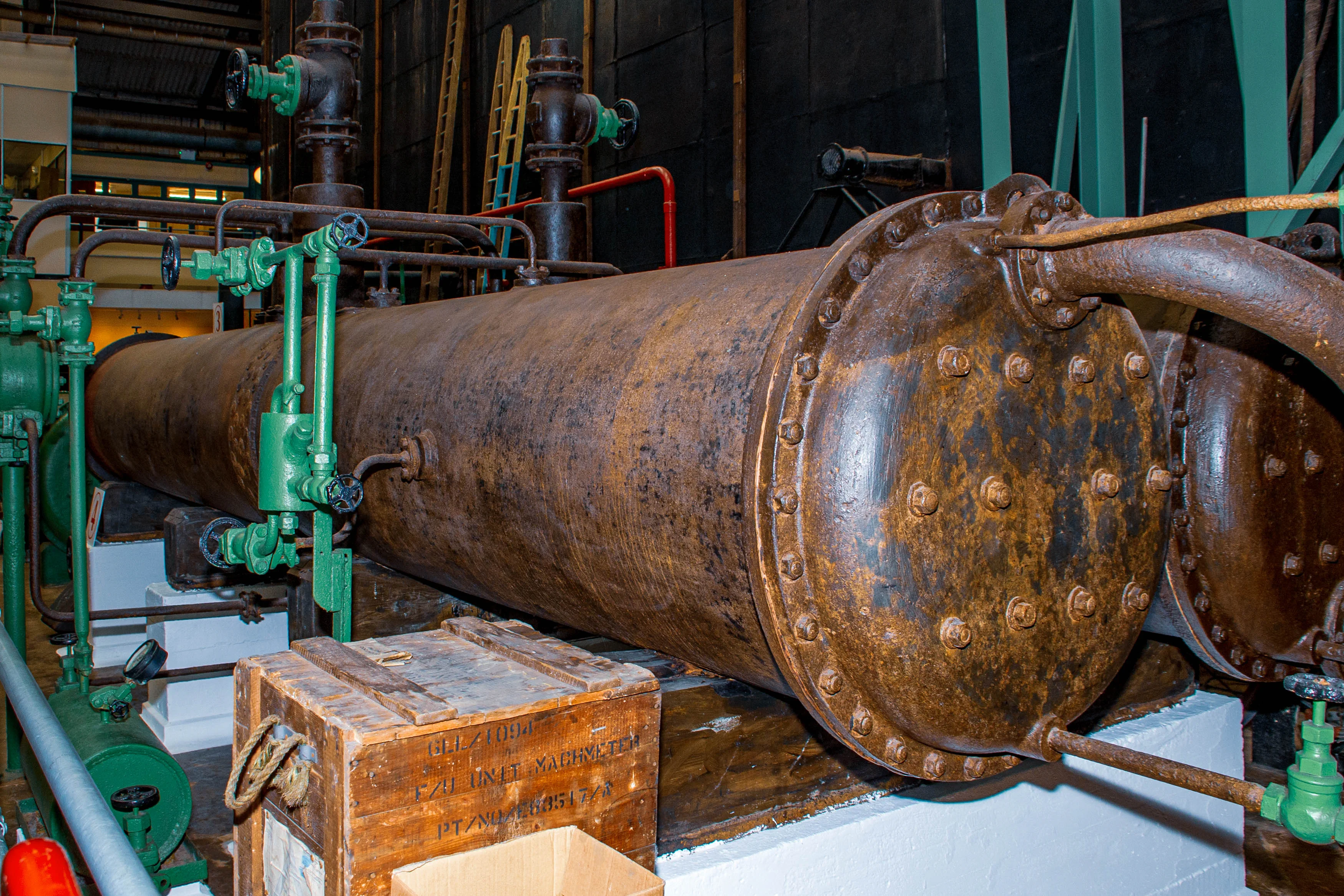 A large, rusty, cylindrical steam boiler with bolted end caps and an intricate system of pipes and valves in an industrial setting. A wooden crate is placed in front of the boiler.