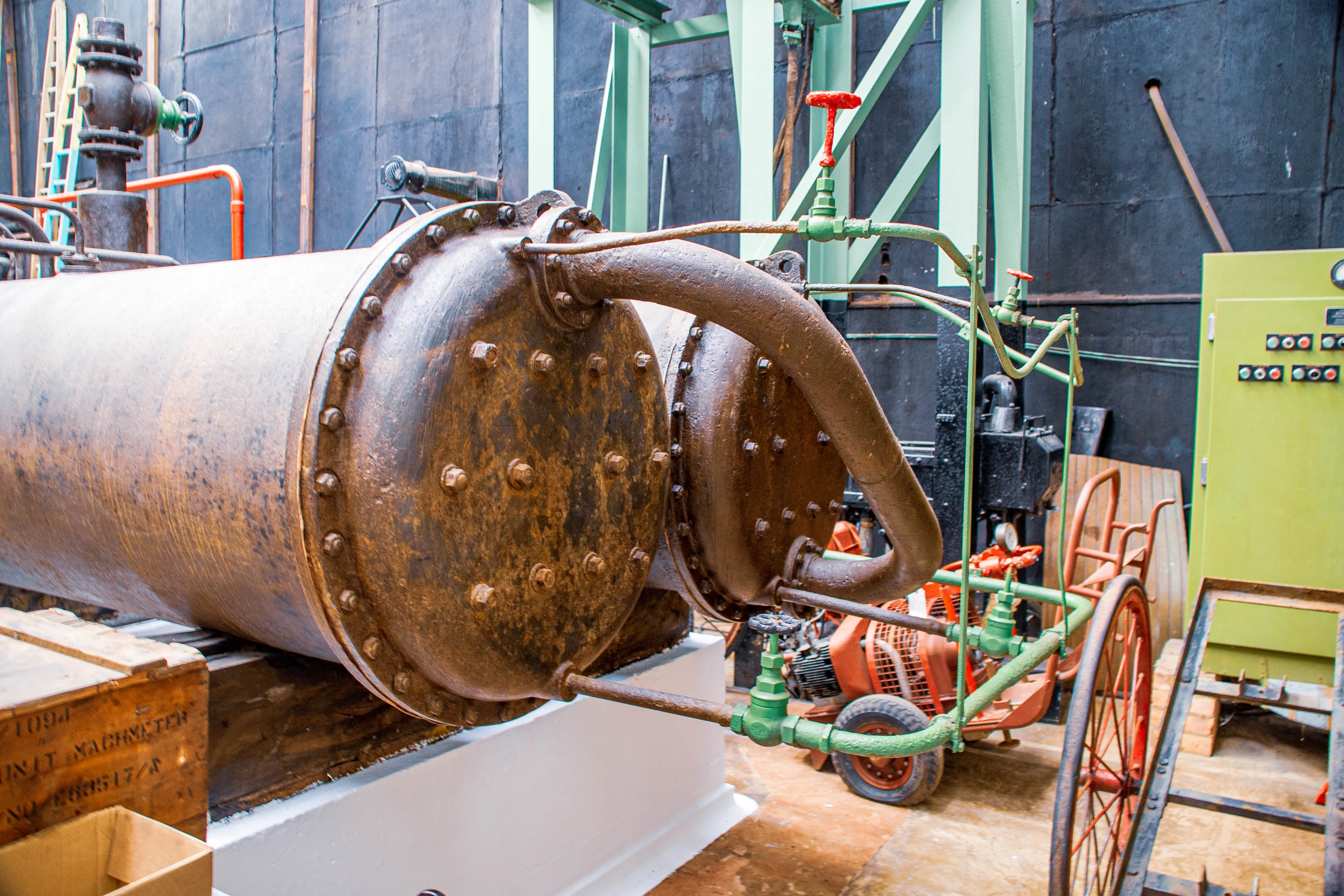 A detailed shot of a large, corroded cylindrical tank with a series of visible bolts around its circumference. Green pipes connect to the tank and lead to other industrial equipment, including a small orange engine with wheels and a green electrical cabinet with buttons.