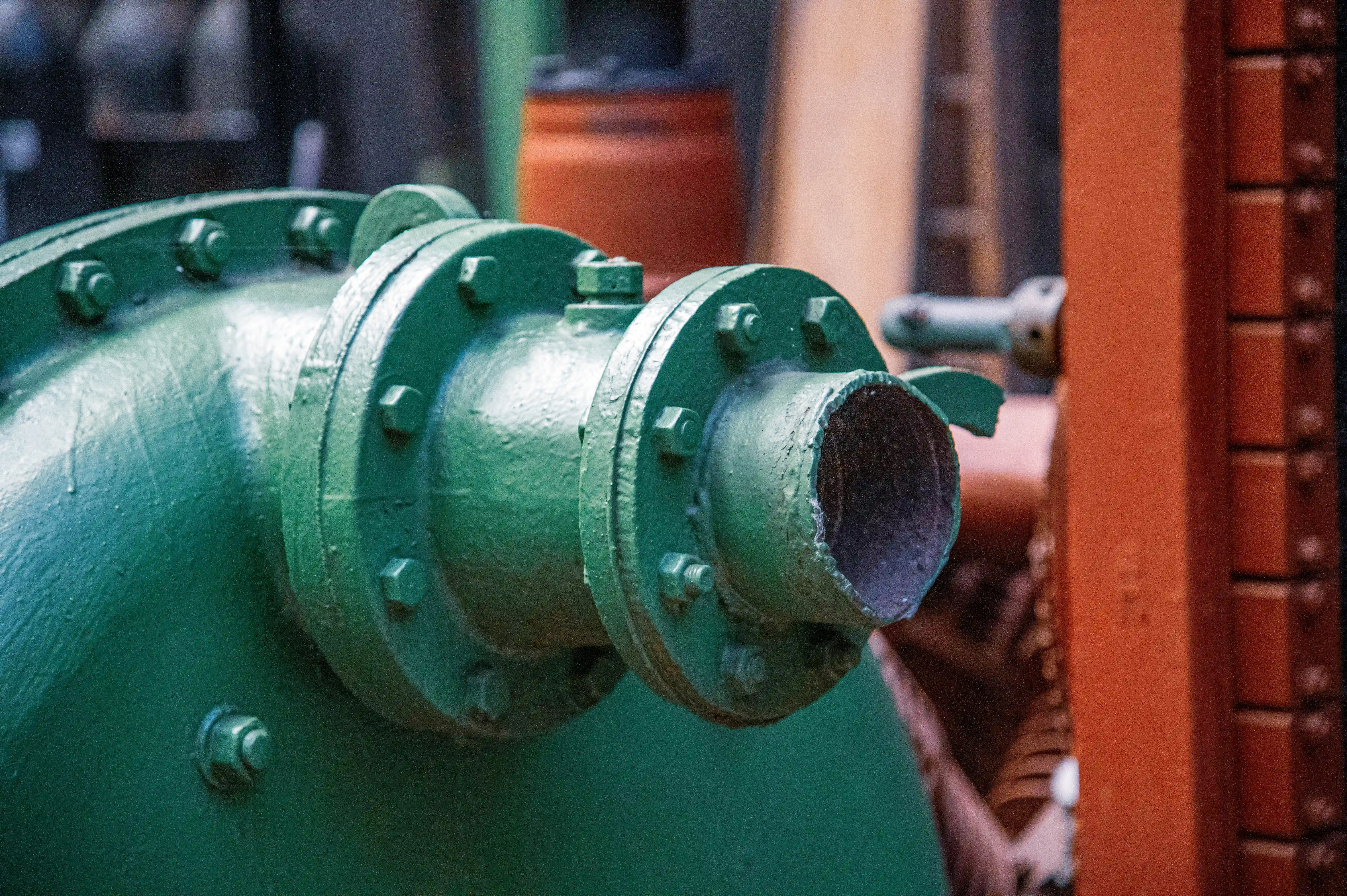 Close-up of a sturdy, green painted metal pipe joint with multiple bolts securing a flange. The end of a connecting pipe is visible and open.