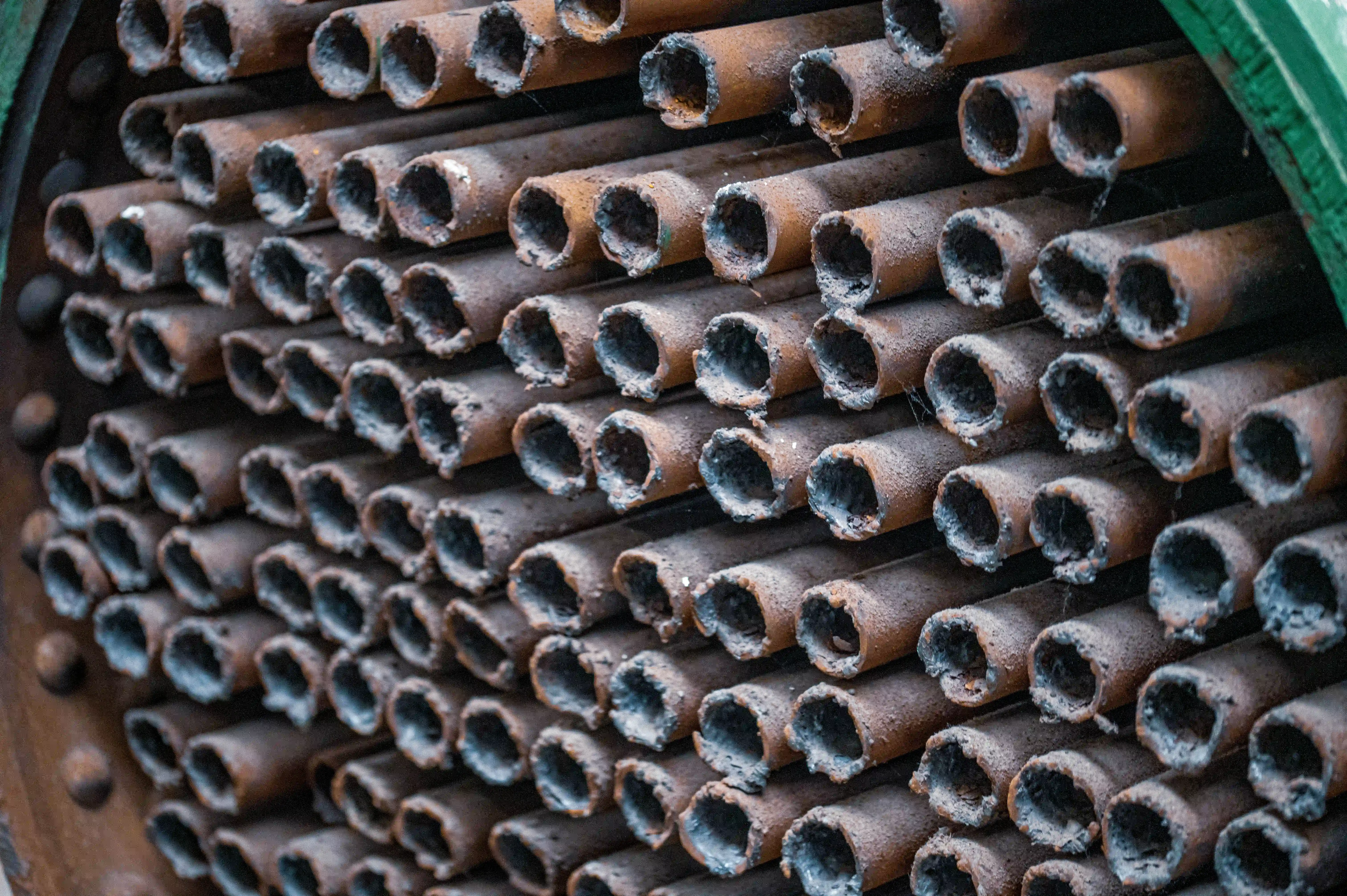Close-up, eye-level shot of a densely packed bank of narrow, cylindrical metal tubes. The tubes are oriented horizontally and arranged in staggered rows, creating a repeating pattern. They are heavily textured with rust, appearing brown and orange, with some areas showing a greyish residue or soot accumulation around the open ends. The interiors of the tubes are dark. On the far right, a sliver of a dark green, metallic edge is visible, likely part of the boiler's casing. The lighting highlights the rough surface of the metal.