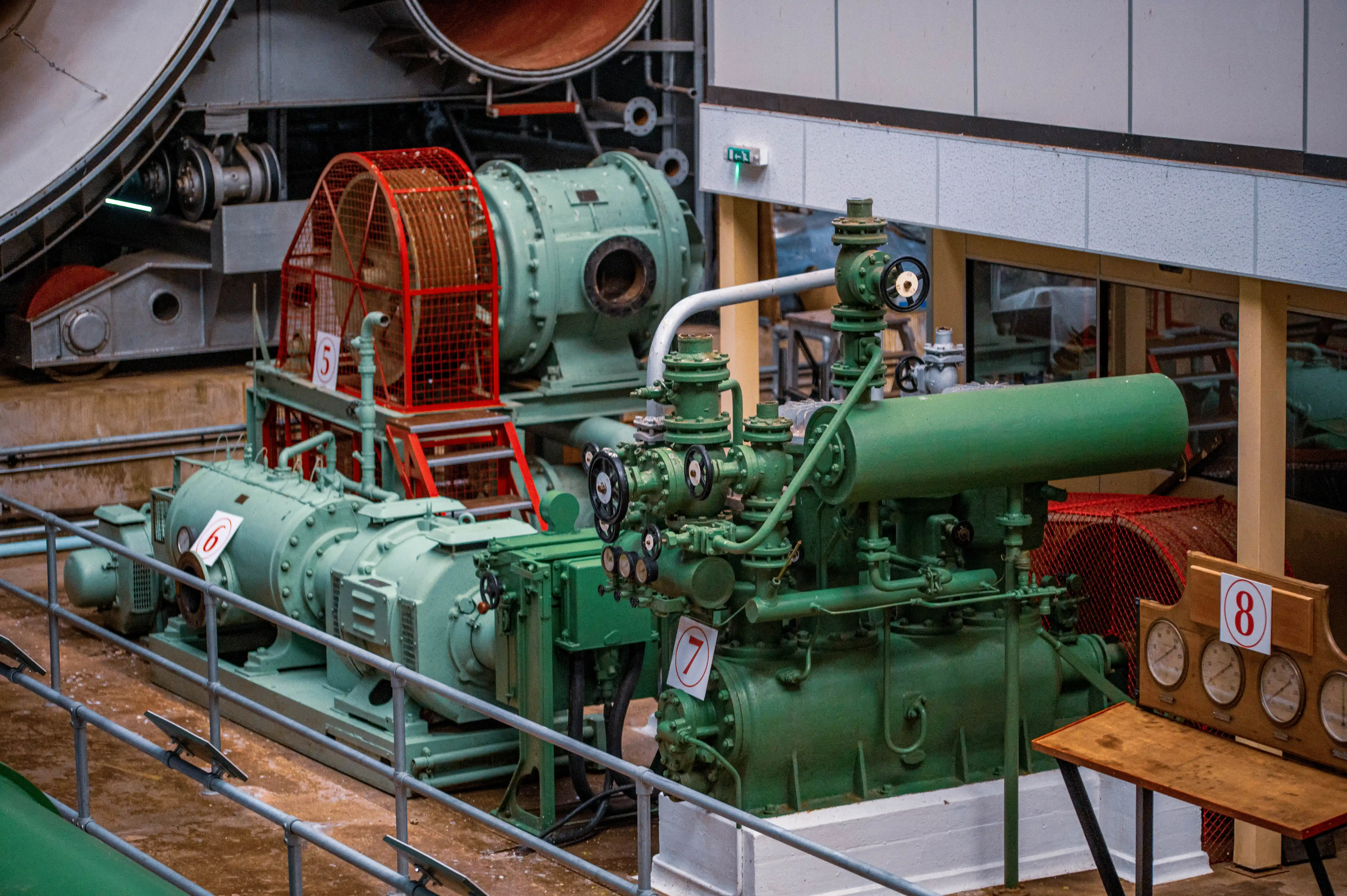 Close-up view of vintage industrial machinery in a dimly lit setting, with prominent green painted components, red safety cages, and various numbered labels and gauges visible.