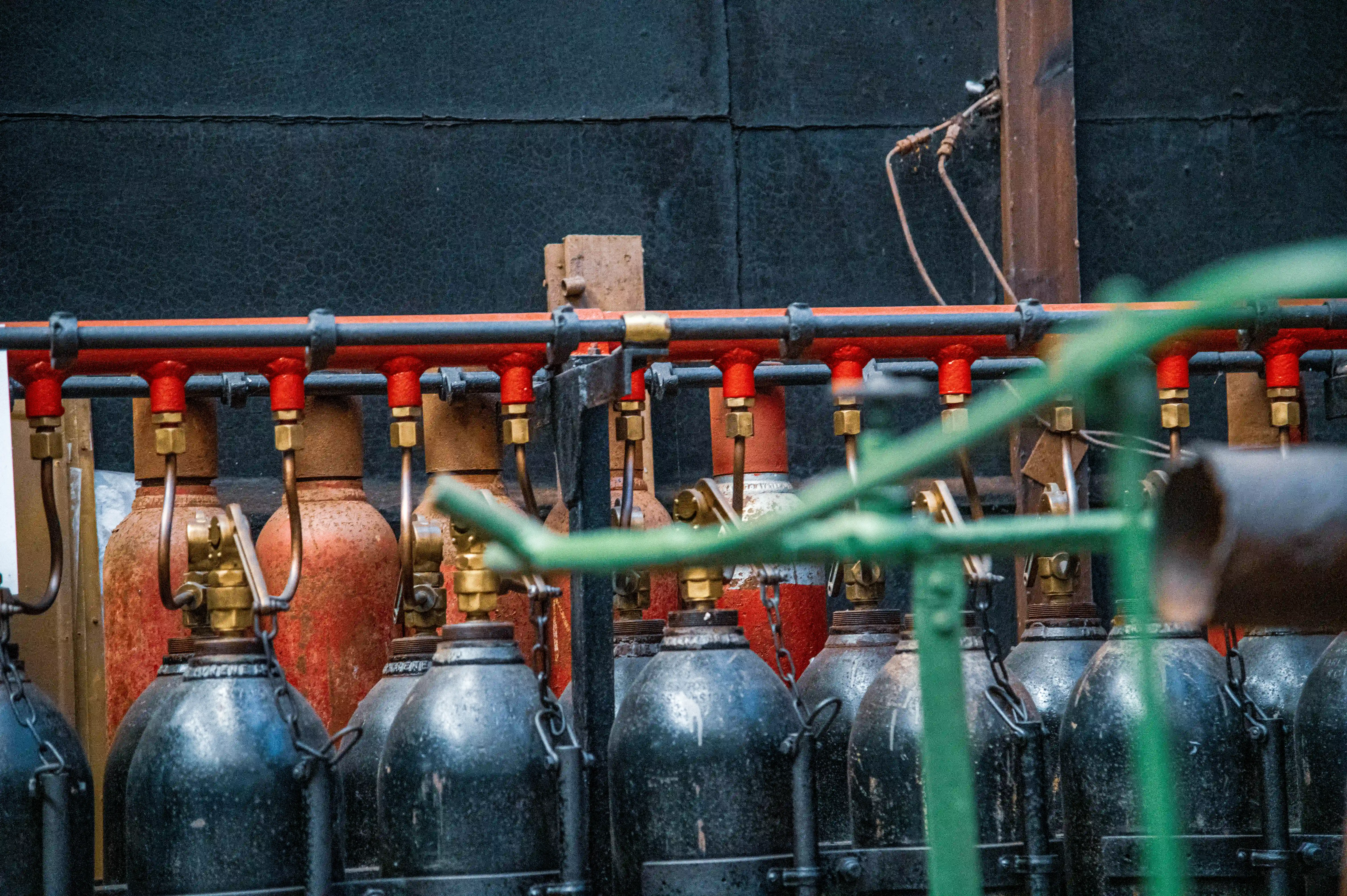  Close-up view of multiple industrial gas cylinders connected to a red distribution manifold with brass fittings and valves, set against a dark, textured wall.