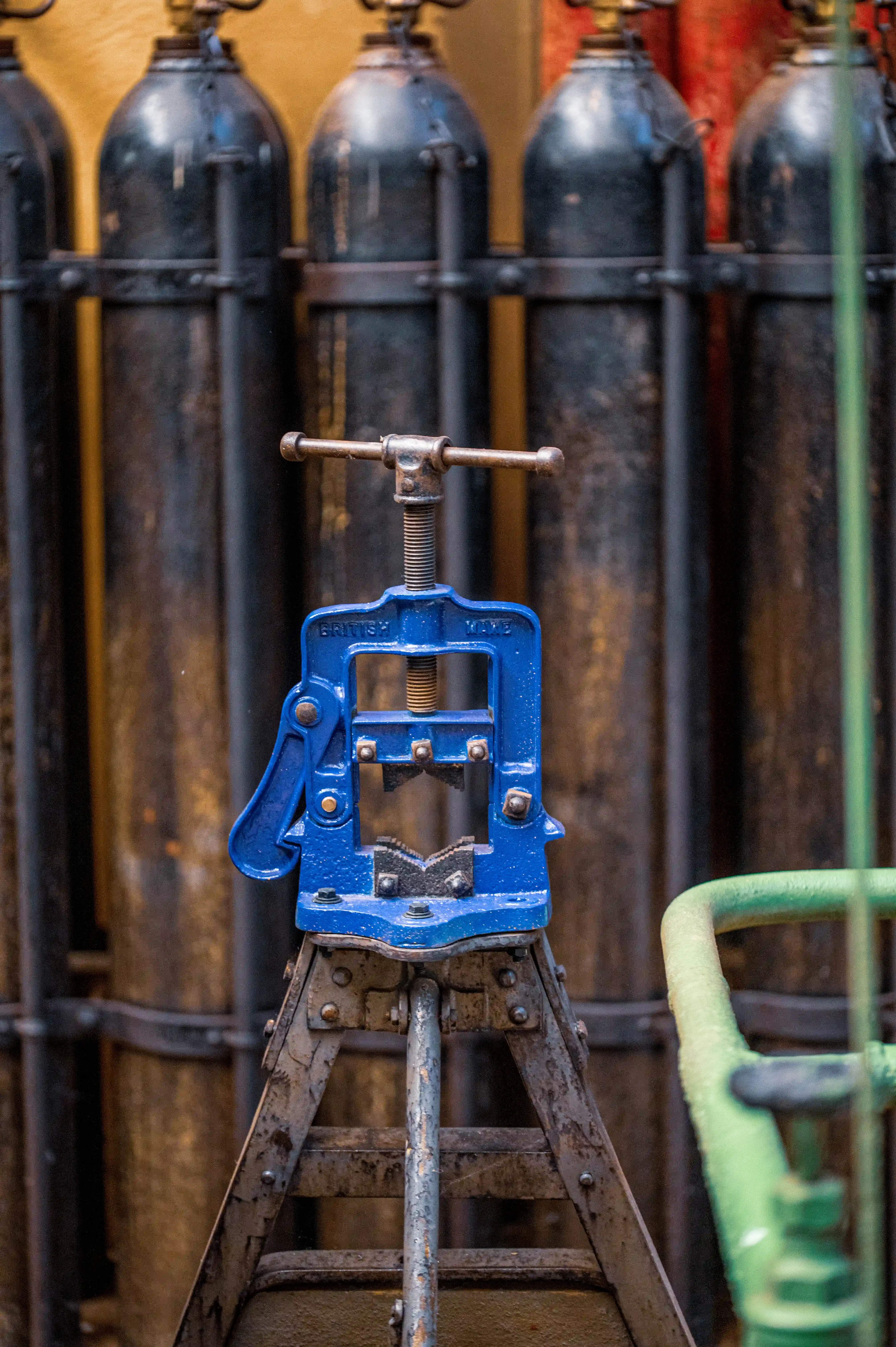 A close-up shot of a blue pipe vise on a metal stand, with out-of-focus industrial gas cylinders in the background.