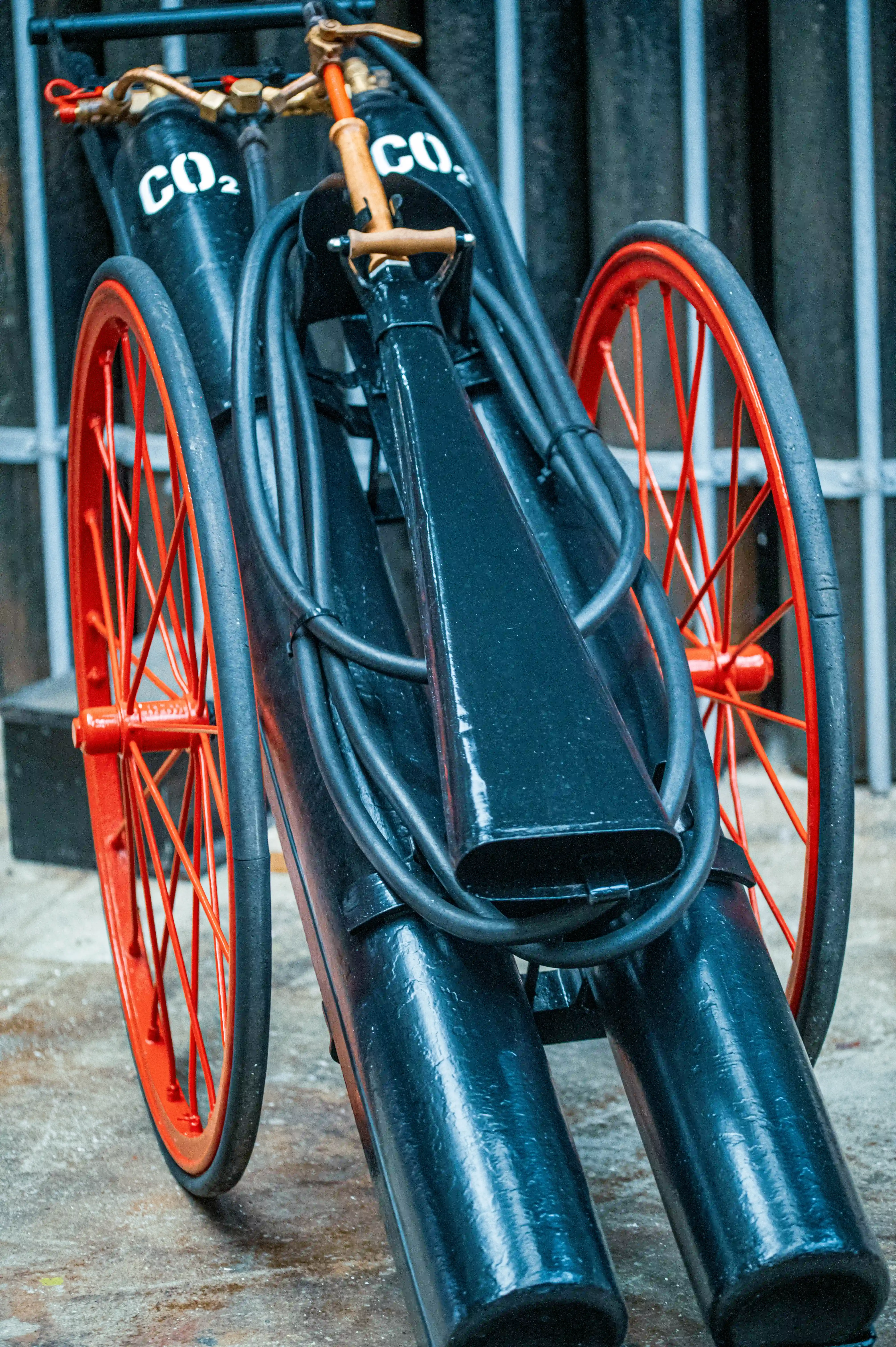 Close-up of a vintage fire extinguisher on red spoked wheels, with two black CO2 cylinders and coiled black hoses.