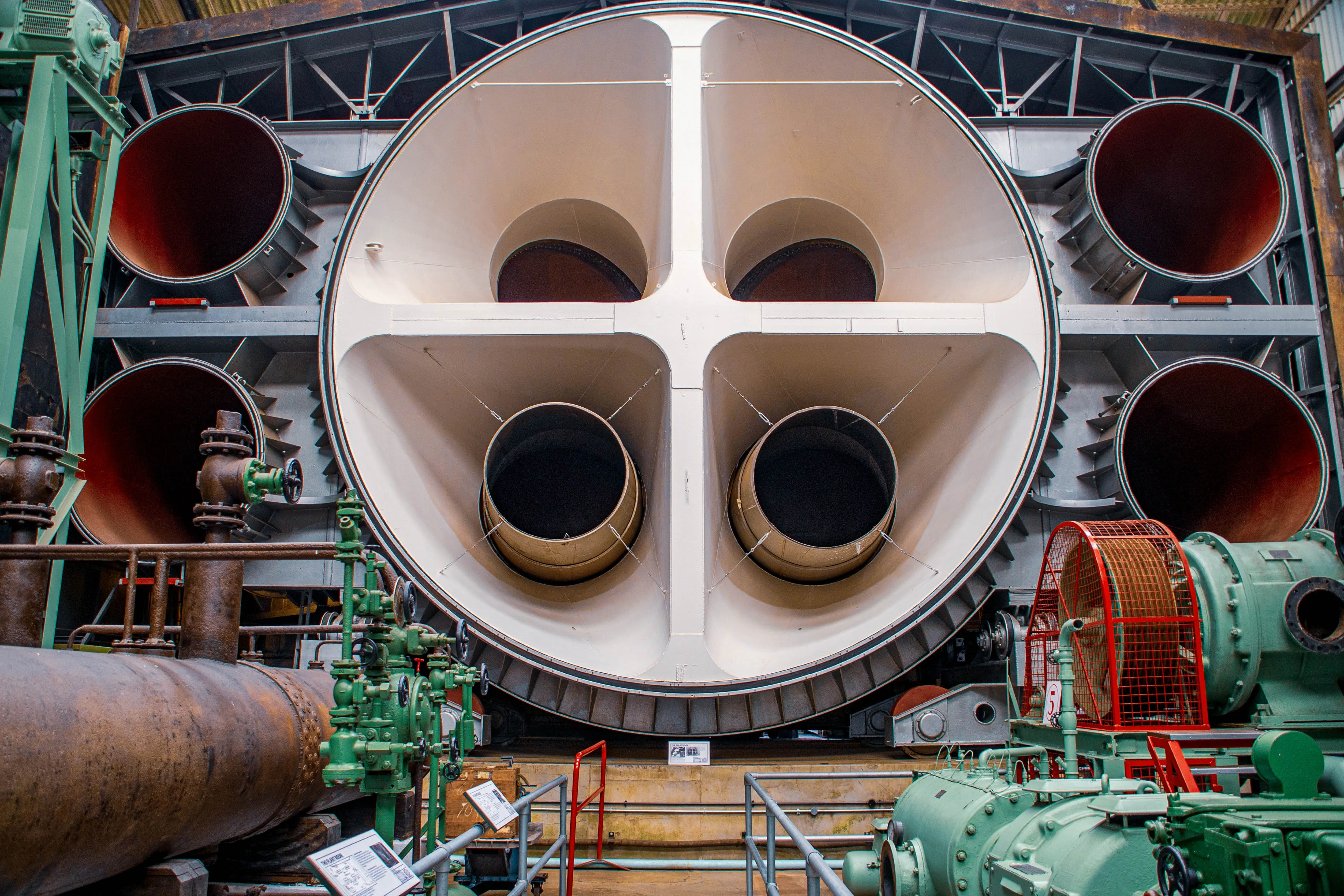 Close-up of a large, complex industrial wind tunnel. A wide, circular aperture dominates the centre, divided into four sections, each leading to a smaller circular opening. Surrounding this are large pipes, green industrial machinery, and metallic structural elements.