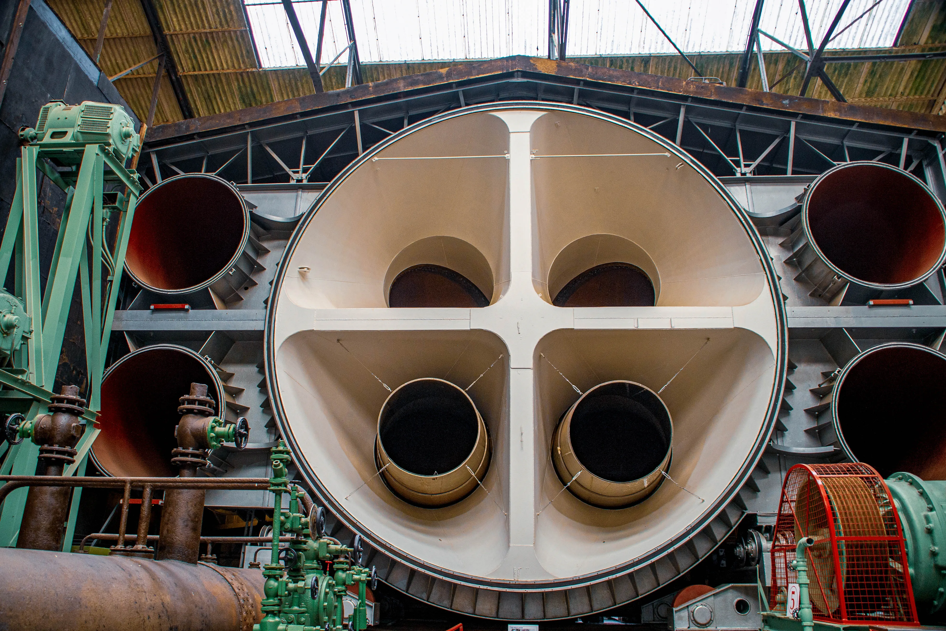 Close-up of a large, complex industrial wind tunnel. A wide, circular aperture dominates the centre, divided into four sections, each leading to a smaller circular opening. Surrounding this are large pipes, green industrial machinery, and metallic structural elements.