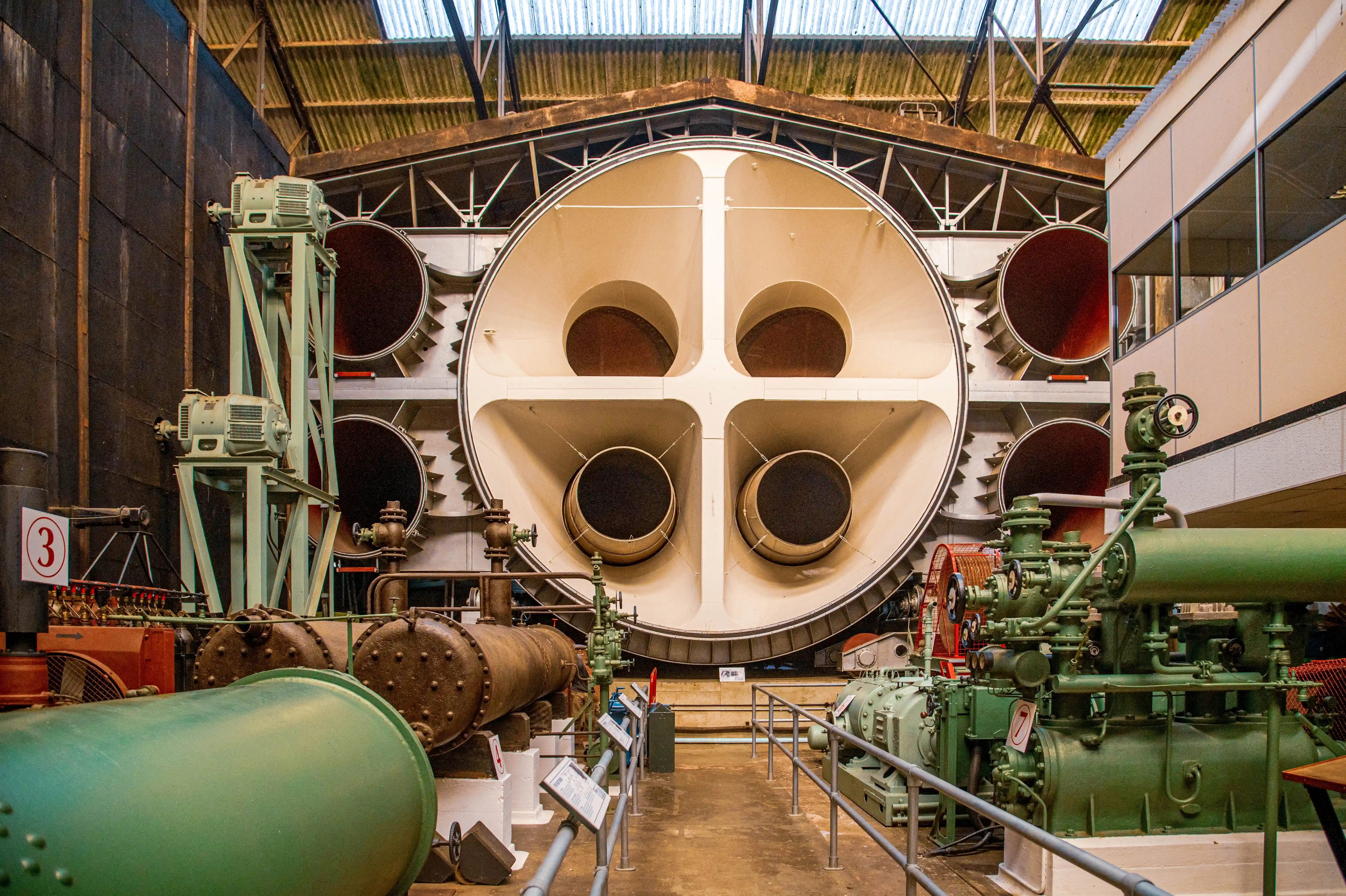 Close-up of a large, complex industrial wind tunnel. A wide, circular aperture dominates the centre, divided into four sections, each leading to a smaller circular opening. Surrounding this are large pipes, green industrial machinery, and metallic structural elements.